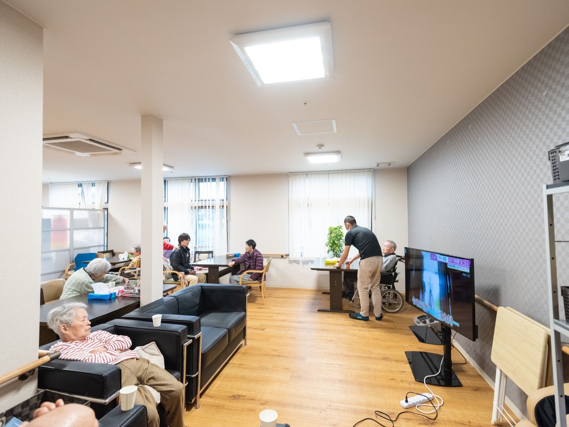 共有の談話スペースに、入居者様が集まっている。室内にはテーブルやソファ、テレビなどが置かれている。壁には手すりが設置されている。