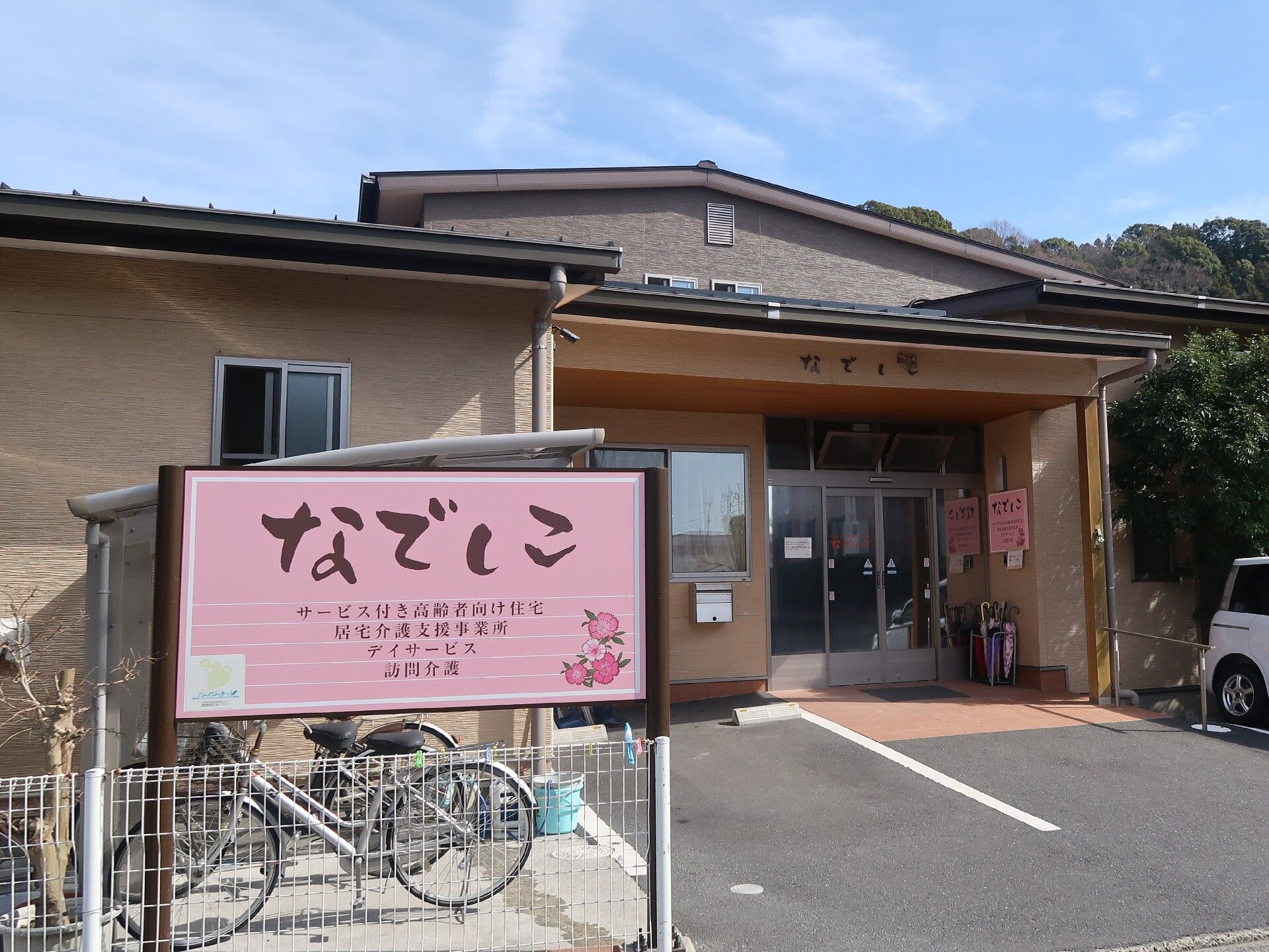 ピンクの看板には住宅名が書かれている。看板の後ろには駐輪場があり、自転車が止められていて、玄関の前には駐車場があり、和風の建物が建っている。