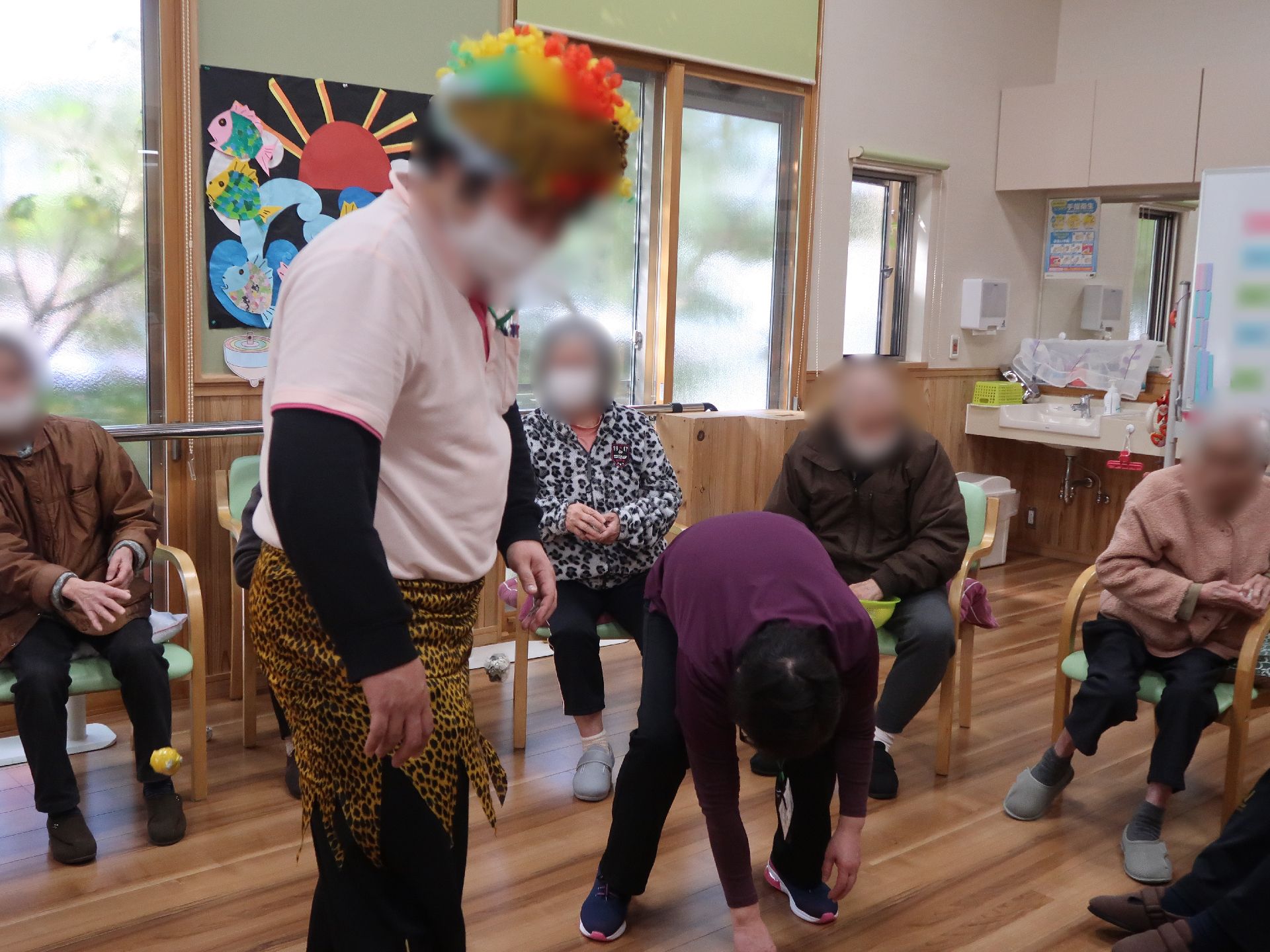 椅子に座った高齢者が周りを囲み、中央にはカラフルなアフロヘアのウィッグをかぶり、動物の皮を使用した風の短パンをはいたスタッフが立っている。