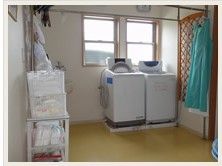 2つの窓のすぐ傍には洗濯機が2台置かれており、一度にまとめて洗濯することができる。白いラックには物が収納されている。