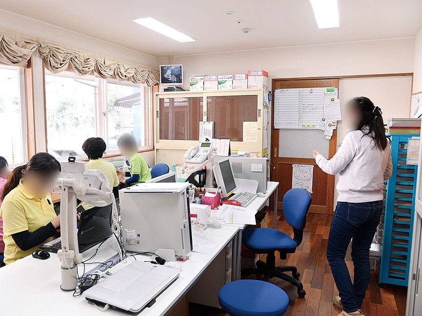 事務机やパソコンなどの並んだ部屋で仕事をするスタッフ達