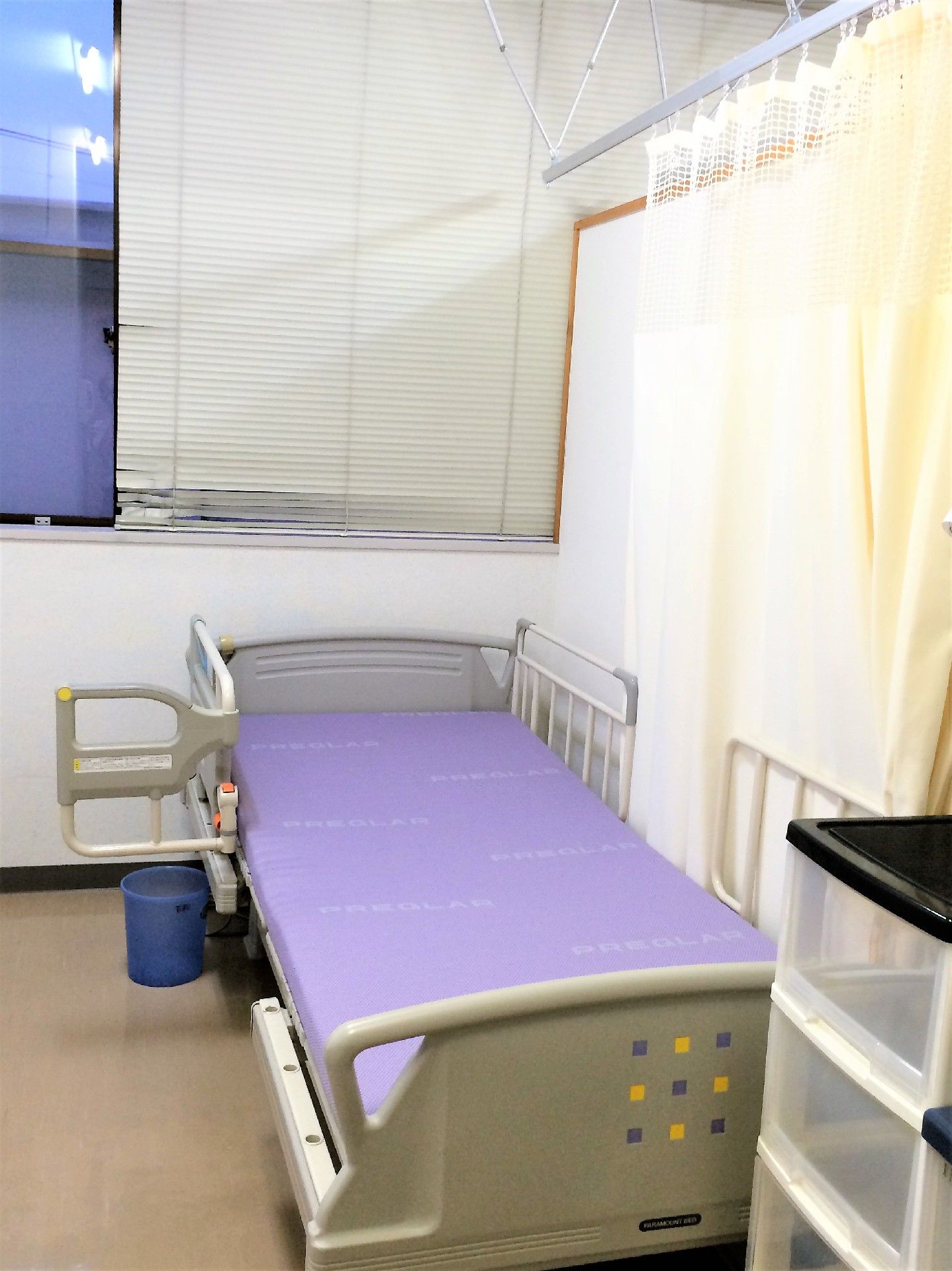 窓にブラインドの付いた居室である。介護用ベッドには、降りたり上がったりする時のサポート役になる取っ手が完備されている。