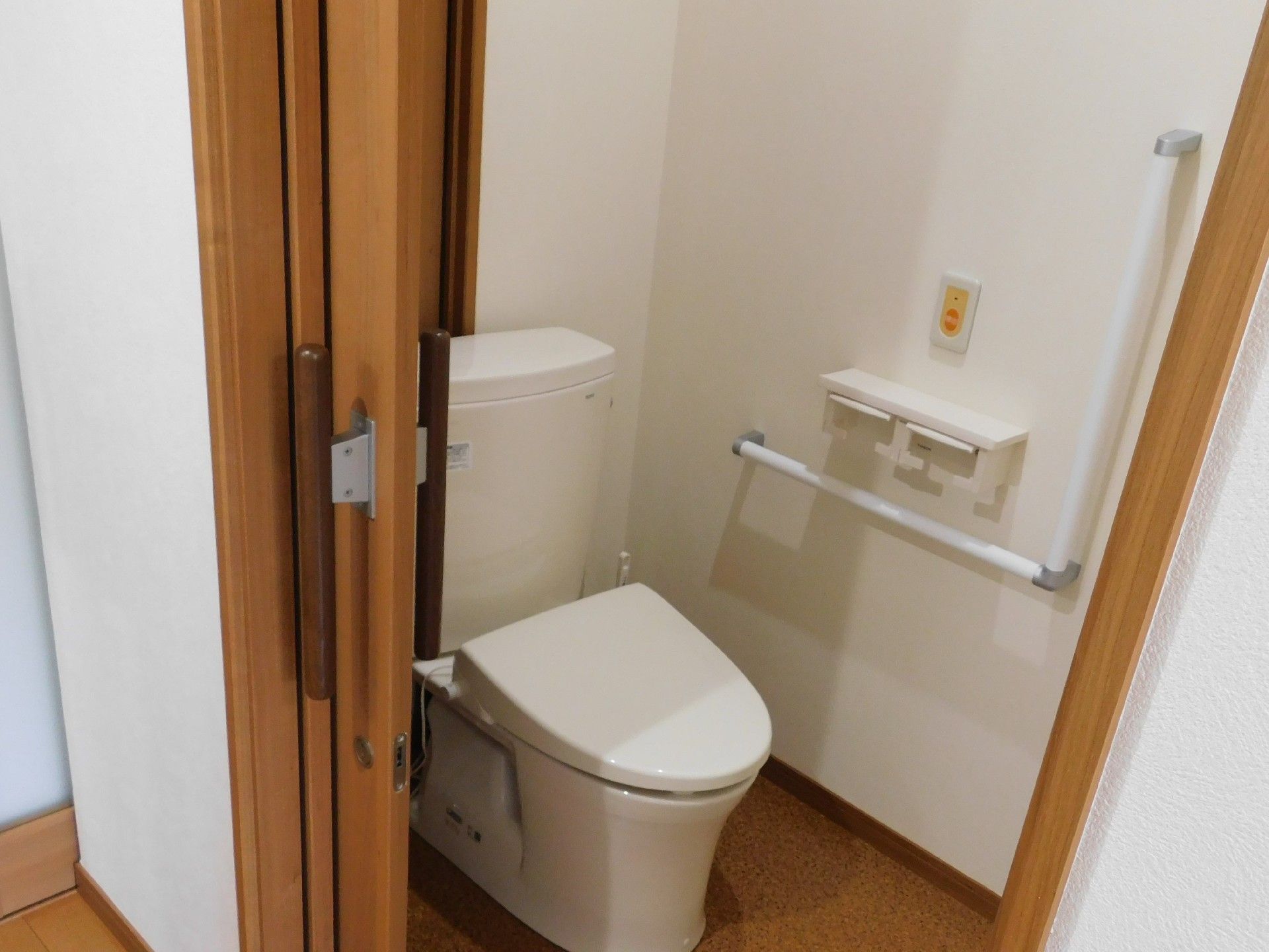 トイレ個室内の様子。壁は白色で、床は茶色系である。洋式便器が1つある。入口の扉は木製で、スライド式になっている。