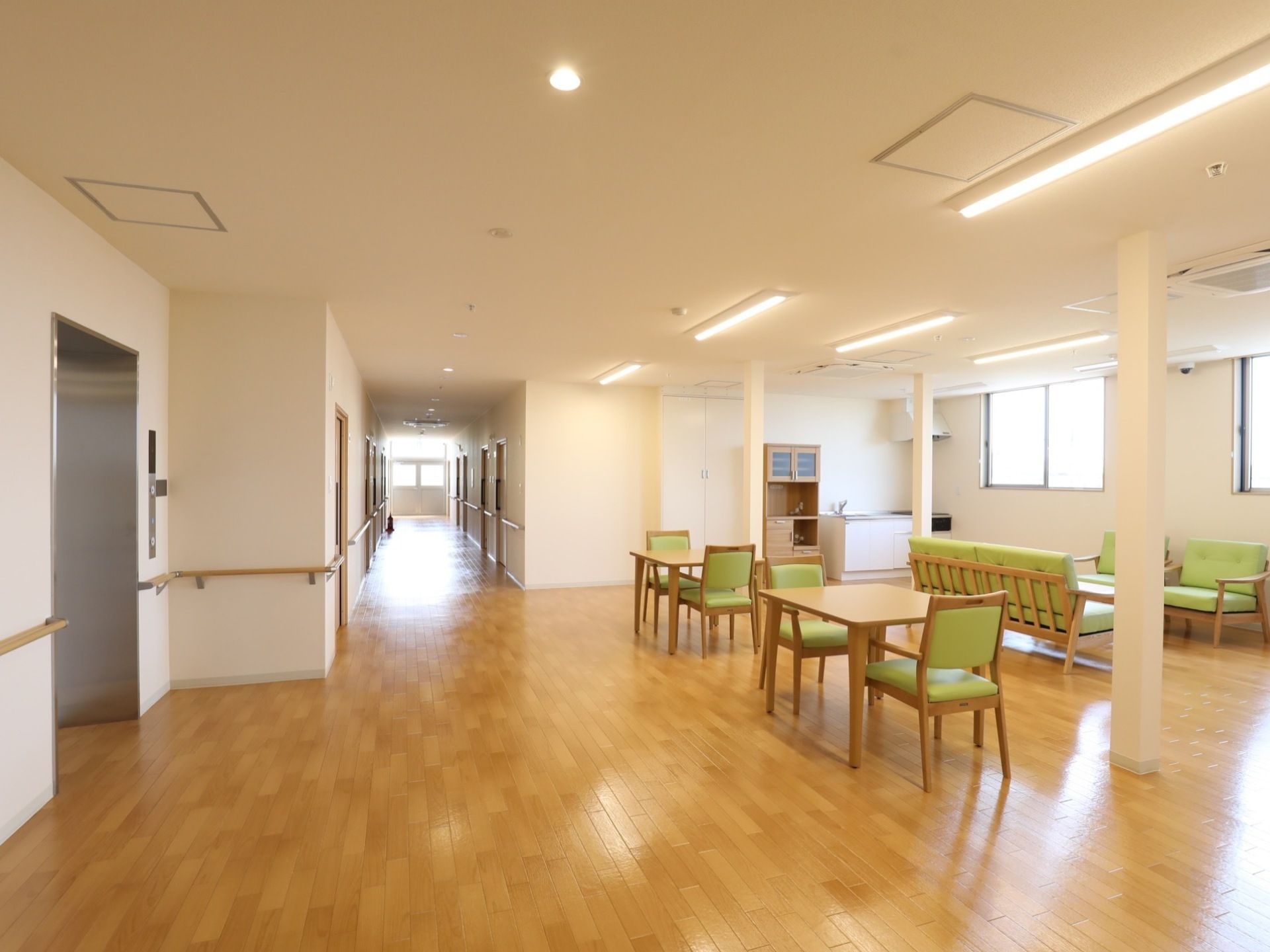 左側の通路から離れた場所に机と椅子がそれぞれ配置されている。椅子のクッション部分はどれも黄緑色をしている。右側奥の壁に台所が設置されている。