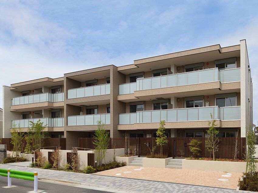 各フロアにベランダがあり、ブラウン系でまとめられた３階建ての建物。階段とスロープがあり、草木も多く植えられている。