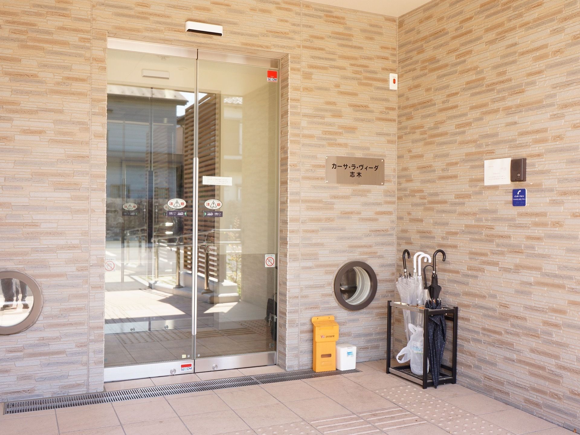 建物入り口の右上には看板、右下には傘立てが設置してある。自動ドアの扉は透明で、中の様子が確認できる。