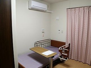 窓に淡いピンク系のカーテンが取り付けられている。ベッドが置かれ、壁にはエアコンと緊急用のナースコールが設置されている。