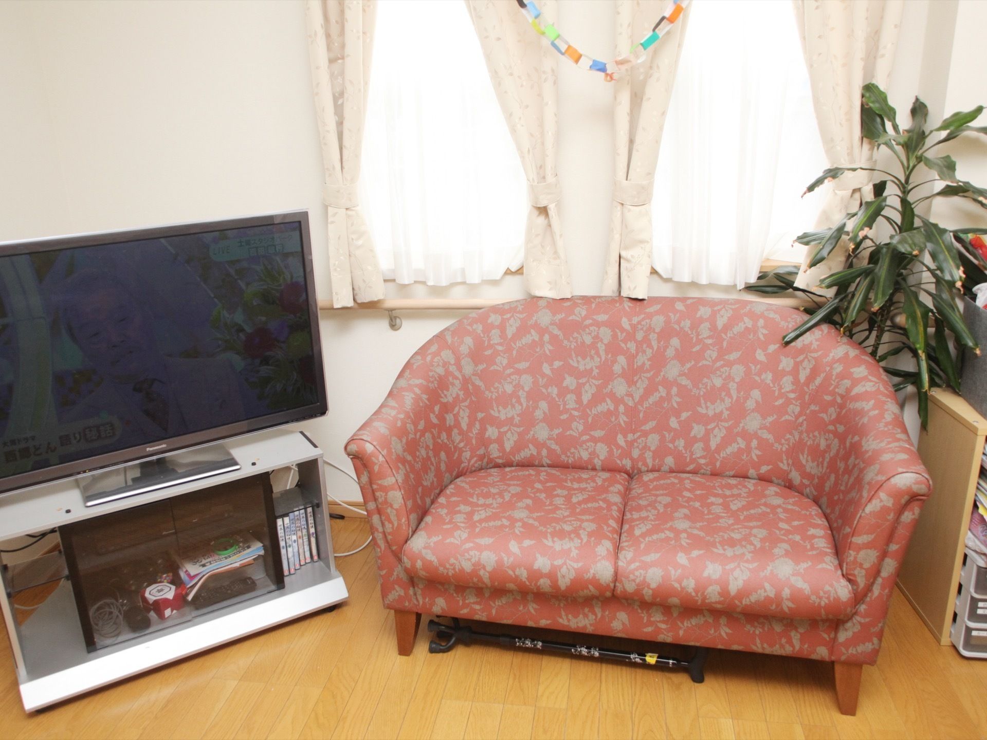 フローリングの床の部屋の窓際には、テレビや赤いソファ、観葉植物やレクリエーションの道具が置かれている。