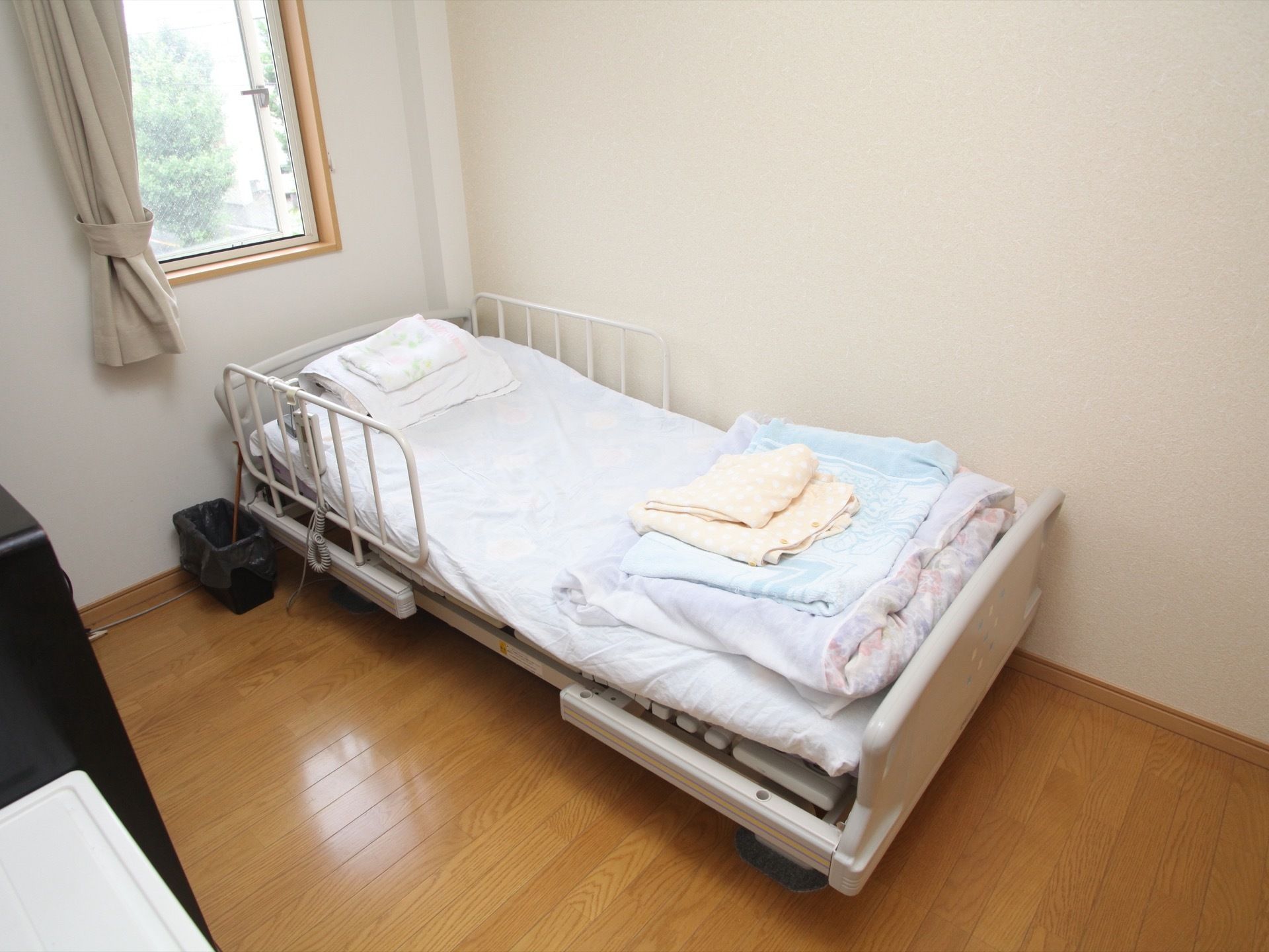 外の景色が見える窓にはカーテンが取り付けられており、その下には介護用ベッドが設置されている。ベッド脇には、ごみ箱と杖が置かれている。