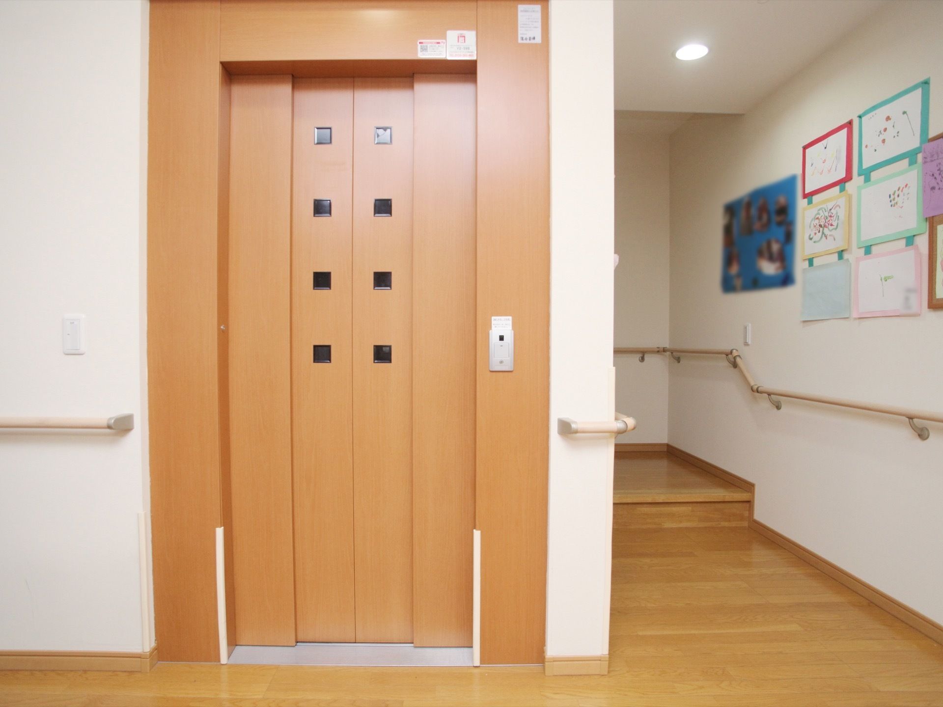 １階の階段横に、木目調の外観のエレベーターが設置されている。壁には手すりがあり、入居者様の作品が掲示されている。