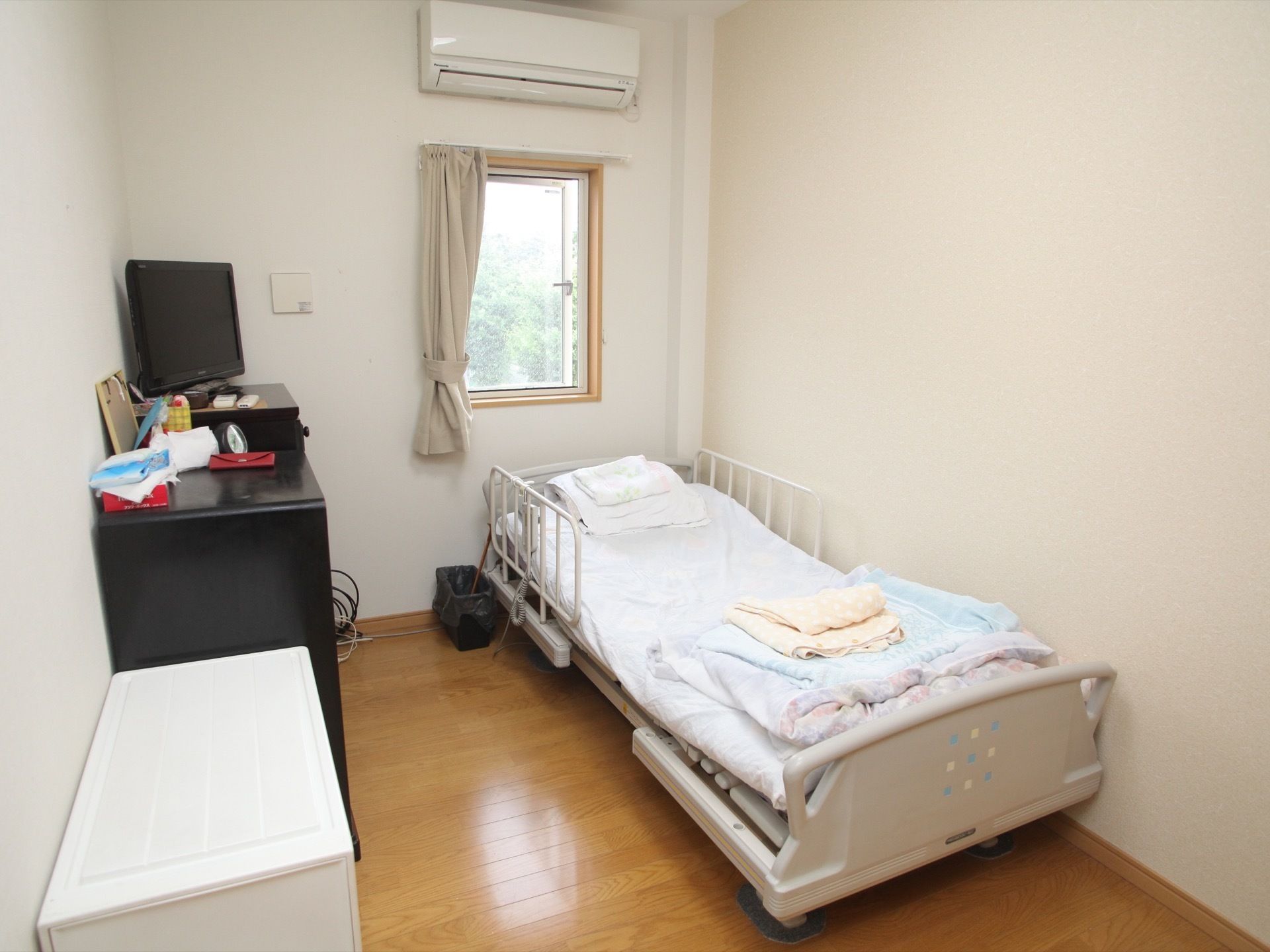 フローリングの部屋に電動ベッドがある。窓が１つあり、その上にはエアコンが設置されている。ベッドの反対側にはチェストとテレビ台もある。