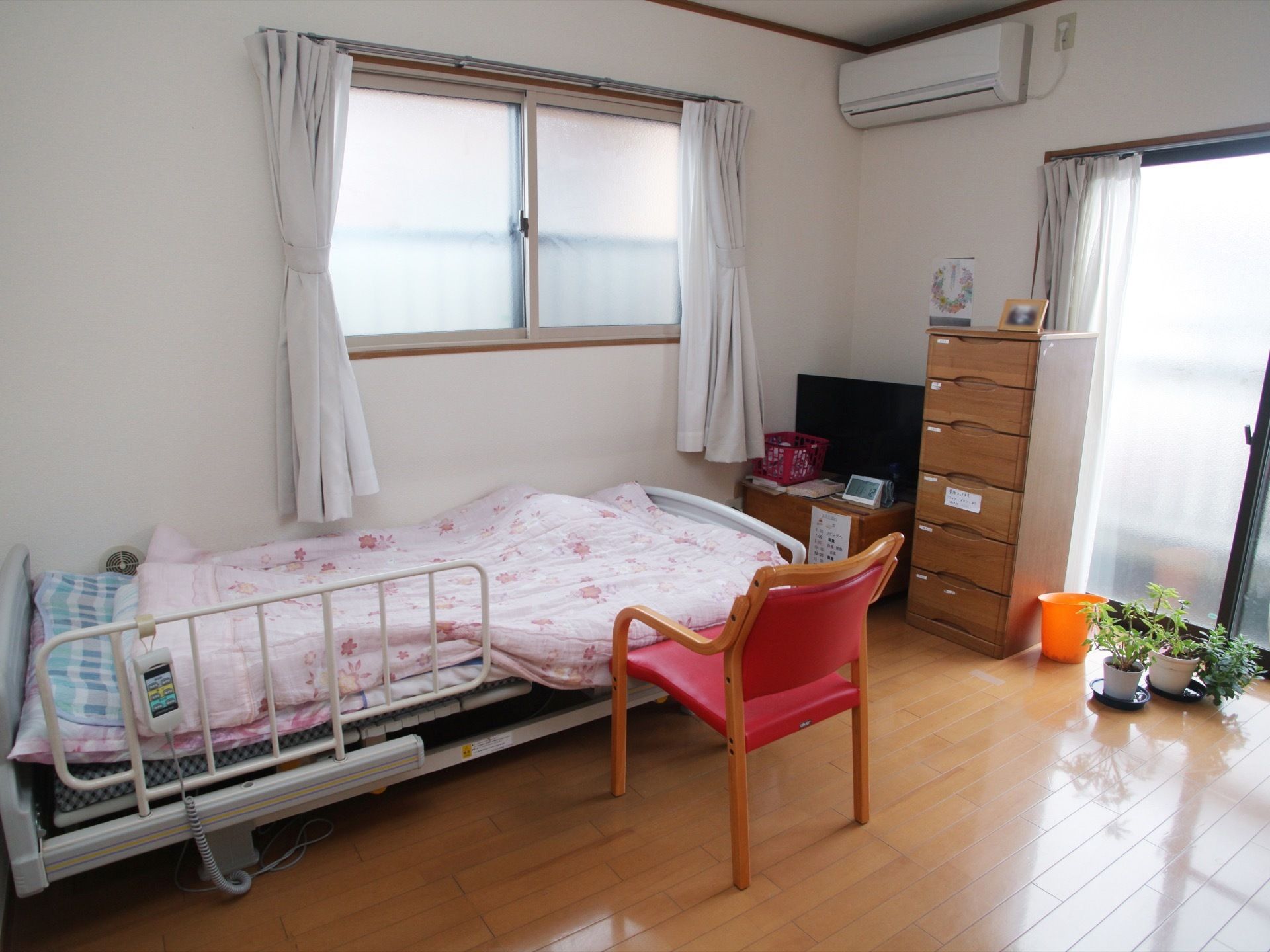 フローリングの部屋に電動ベッドが置かれている。タンスとテレビがあり、壁にはエアコンが設置されている。