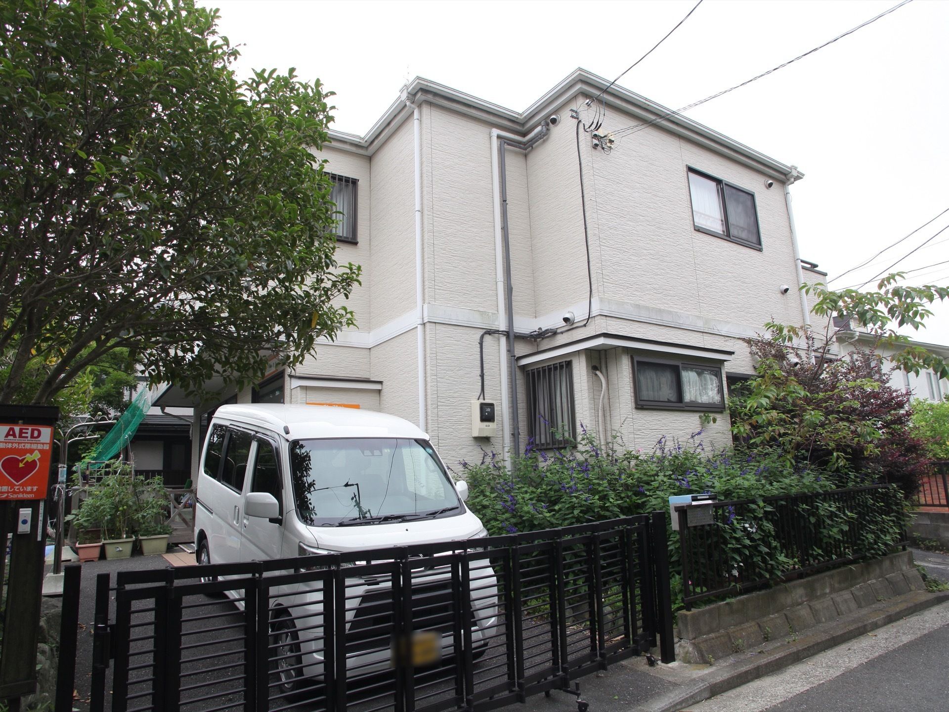 ホームは２階建ての一軒家になっている。壁は明るい灰色で落ち着いた雰囲気である。白い車が駐車スペースに停まっている。
