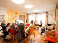 リビングに入居者様が集っており、スタッフと談話している。壁にはホワイトボードやカレンダーなどが貼られている。