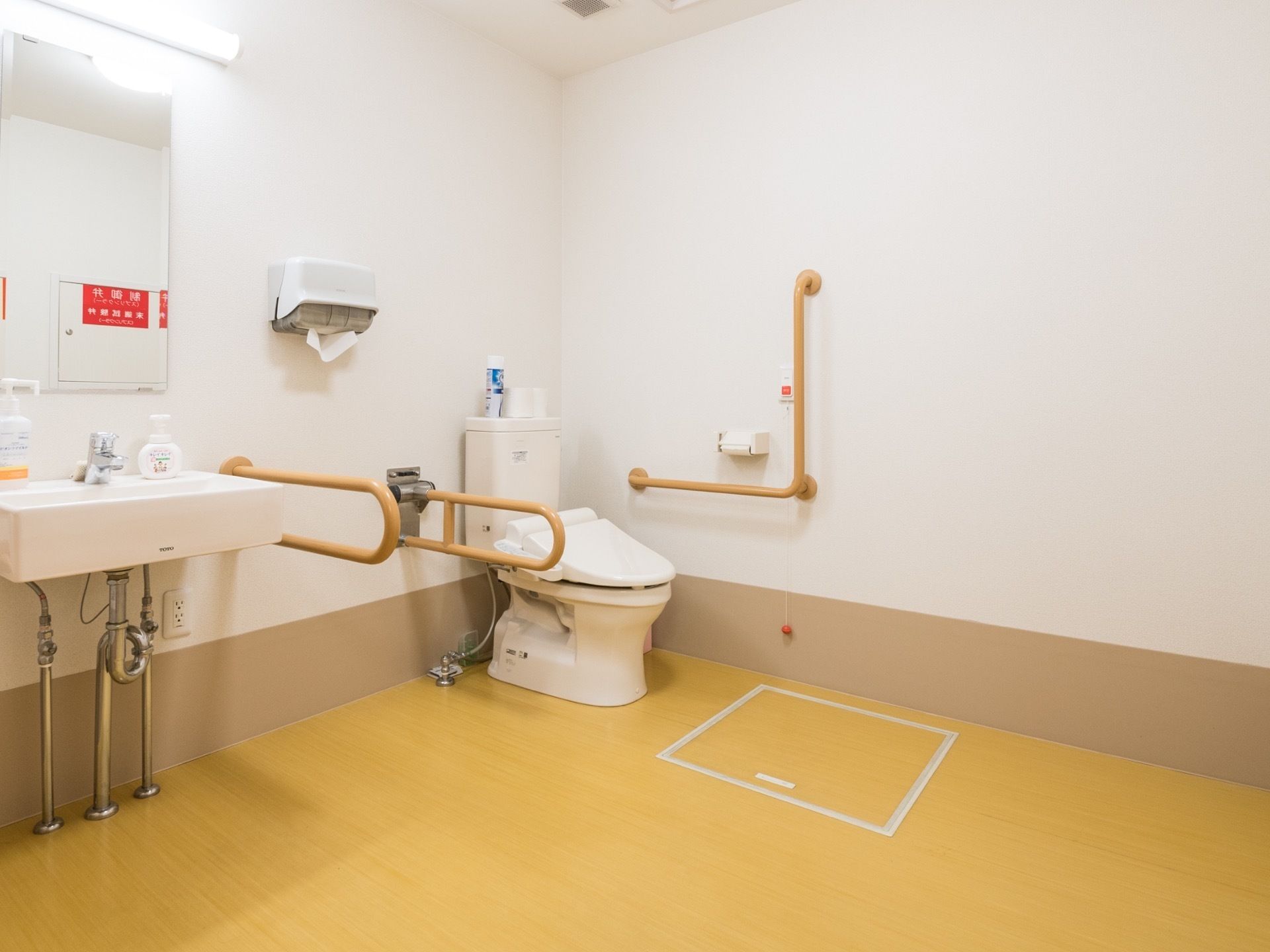 白基調で広い空間のトイレは、壁にはL字型の手すり、手洗い洗面台、トイレットペーパーホルダー、ナースコールが設置されている。
