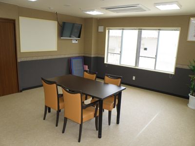 ゆとりのある広いスペースで、部屋の中央に４人掛けの黒とオレンジの椅子と黒いテーブルが設置されている。壁には大型のテレビが備え付けられている。