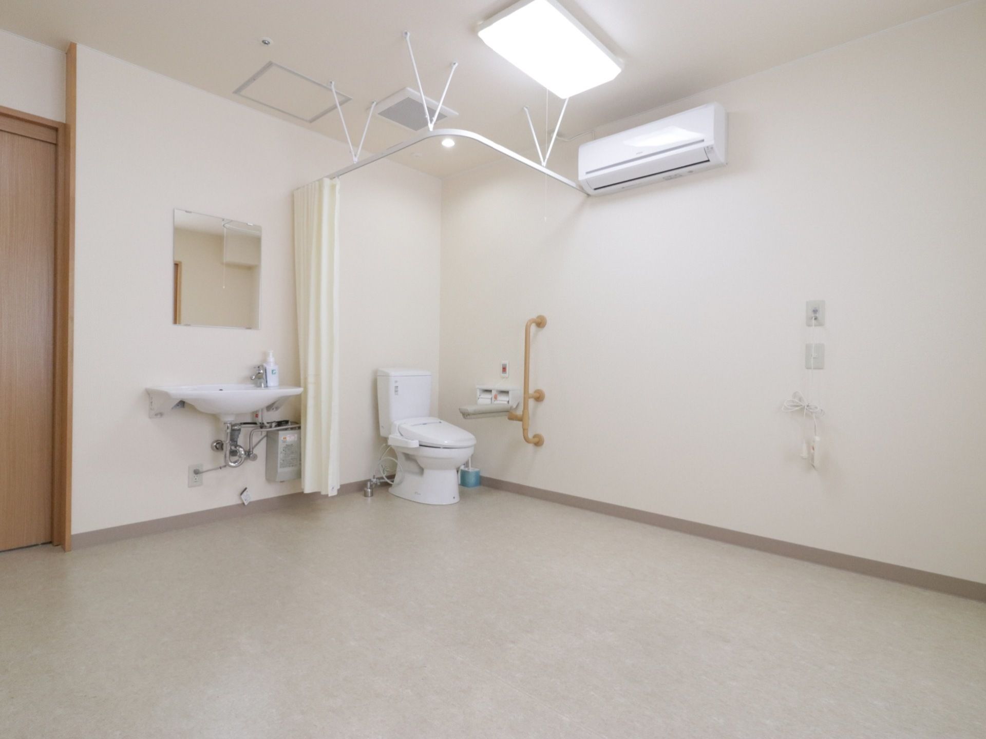 広い部屋の入口横に洗面台が設置されている。レールカーテンで仕切られたスペースにトイレがあり、壁にエアコンと非常ボタンがある。