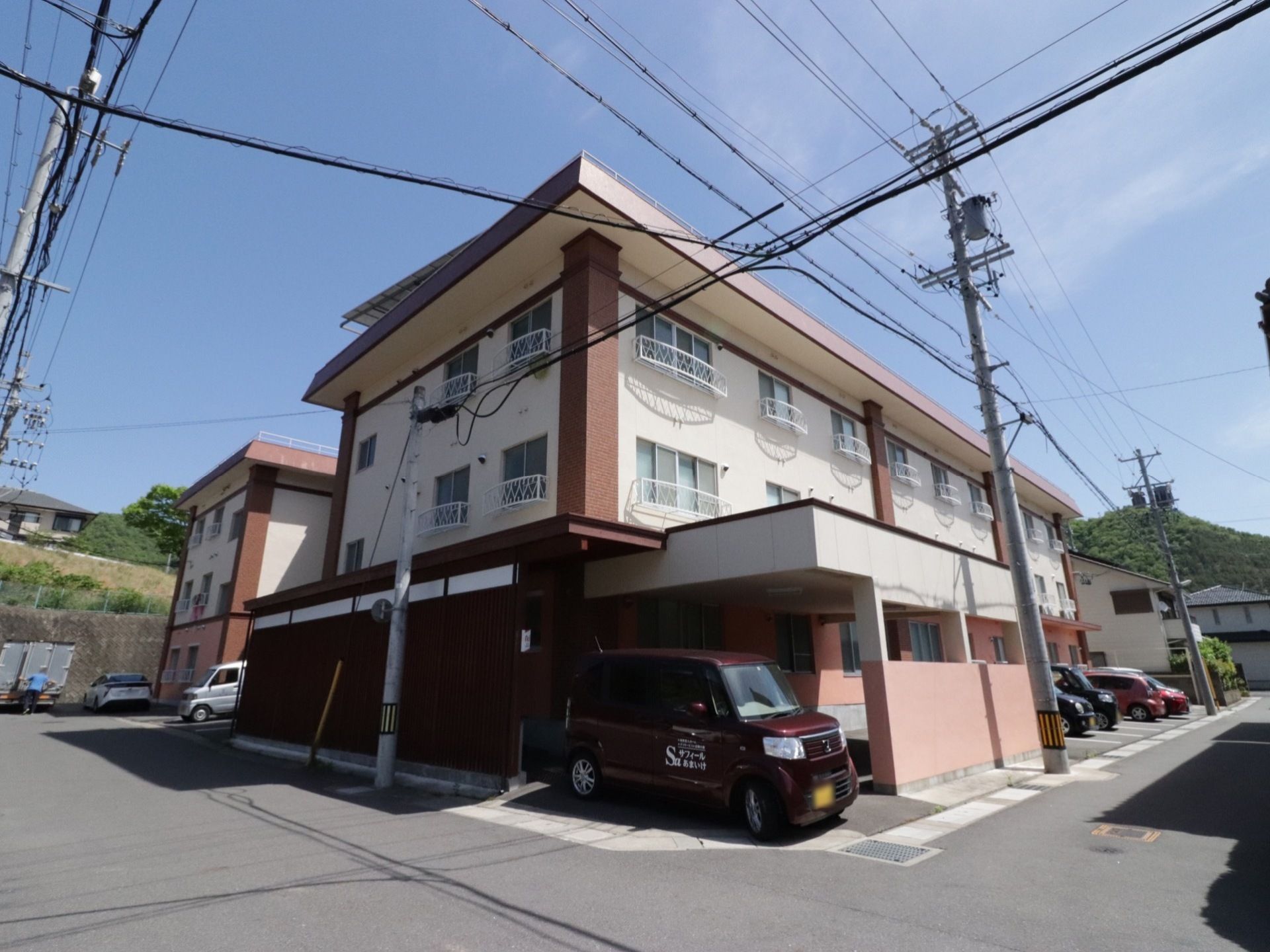 ３階建ての建物で、敷地内に施設所有の軽自動車が１台置かれている。建物に沿って駐車スペースが並んでいる。
