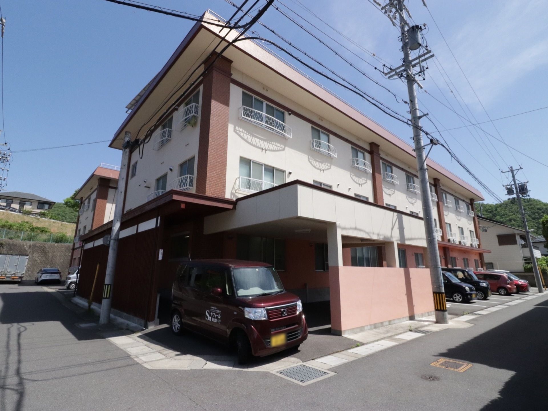 施設は路地の角地に建てられており、各部屋の窓には柵が設置されている。玄関前に施設の車両が止まっている。