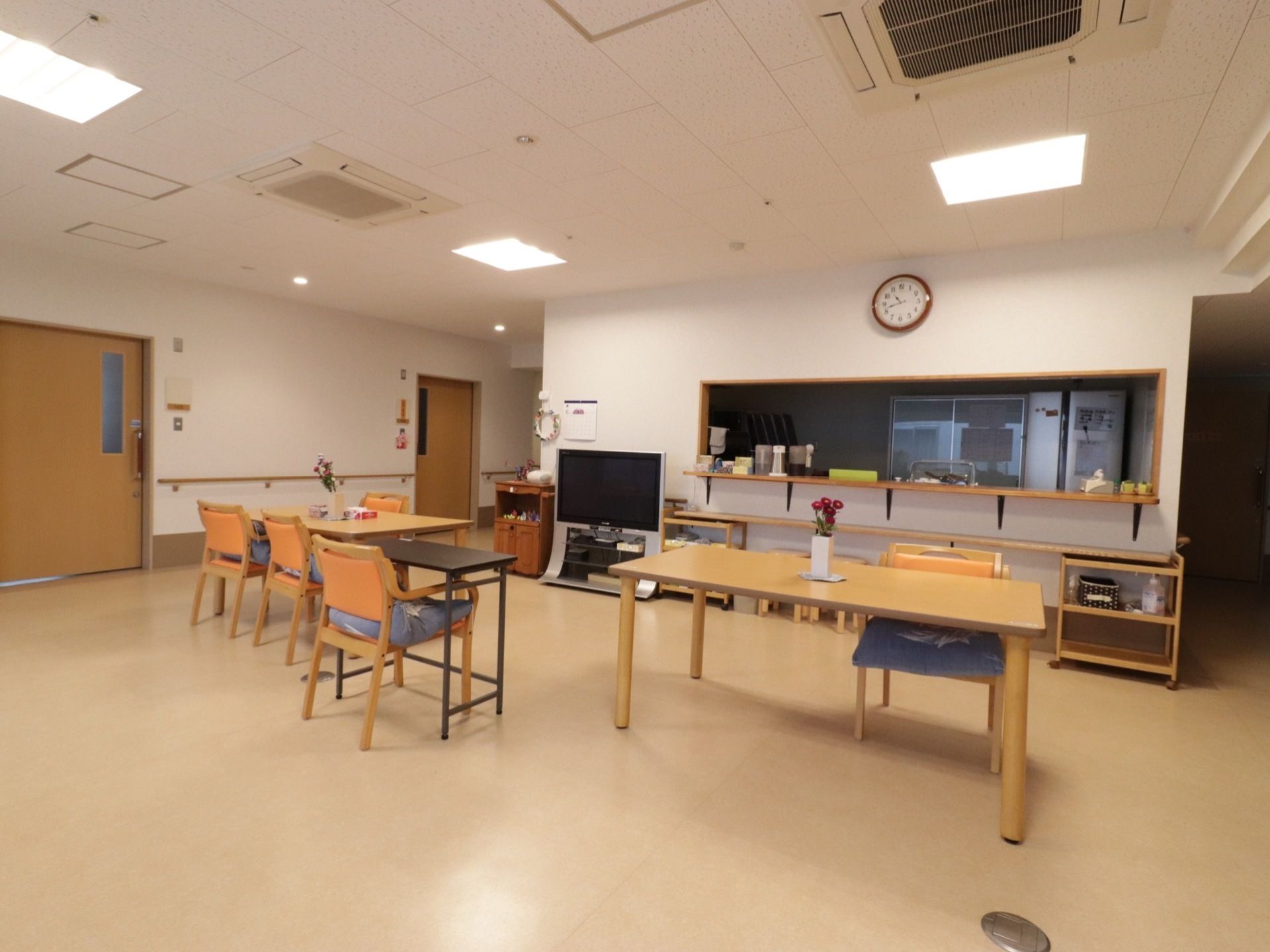 広い室内に複数のテーブルと椅子が置かれ、近くに大型テレビがある。また、奥には対面式のキッチンがある。