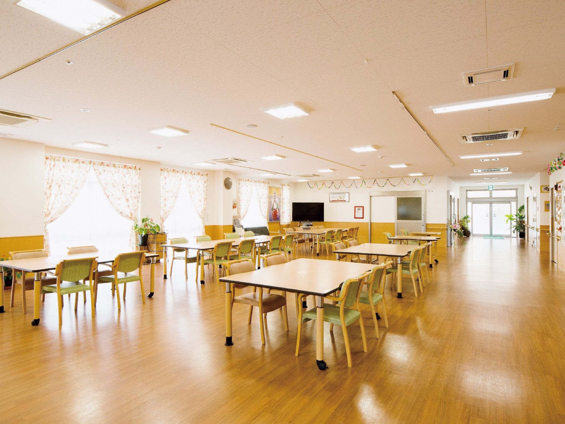 広々とした高齢者施設の食堂。木製のテーブルと緑の椅子が複数配置されている。