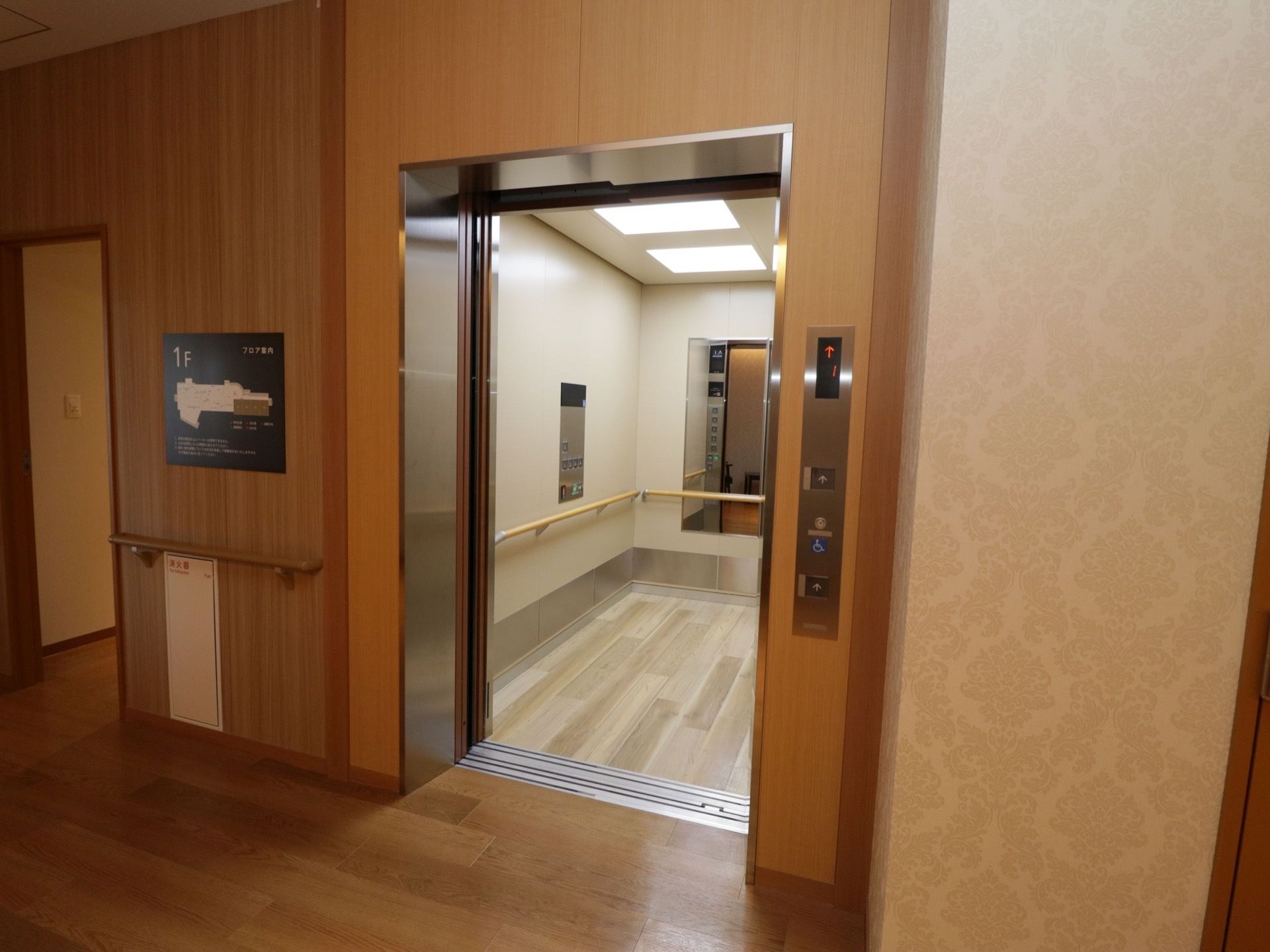 木目調の壁に囲まれたエレベーターで、扉が開いている。扉の左側の壁にフロアマップと手すりがある。右側に呼出ボタンがある。