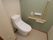 緑と白の壁で囲まれたトイレで、最新型のタイプの温水洗浄便座が取りつけられている。木目調の手すりがある。