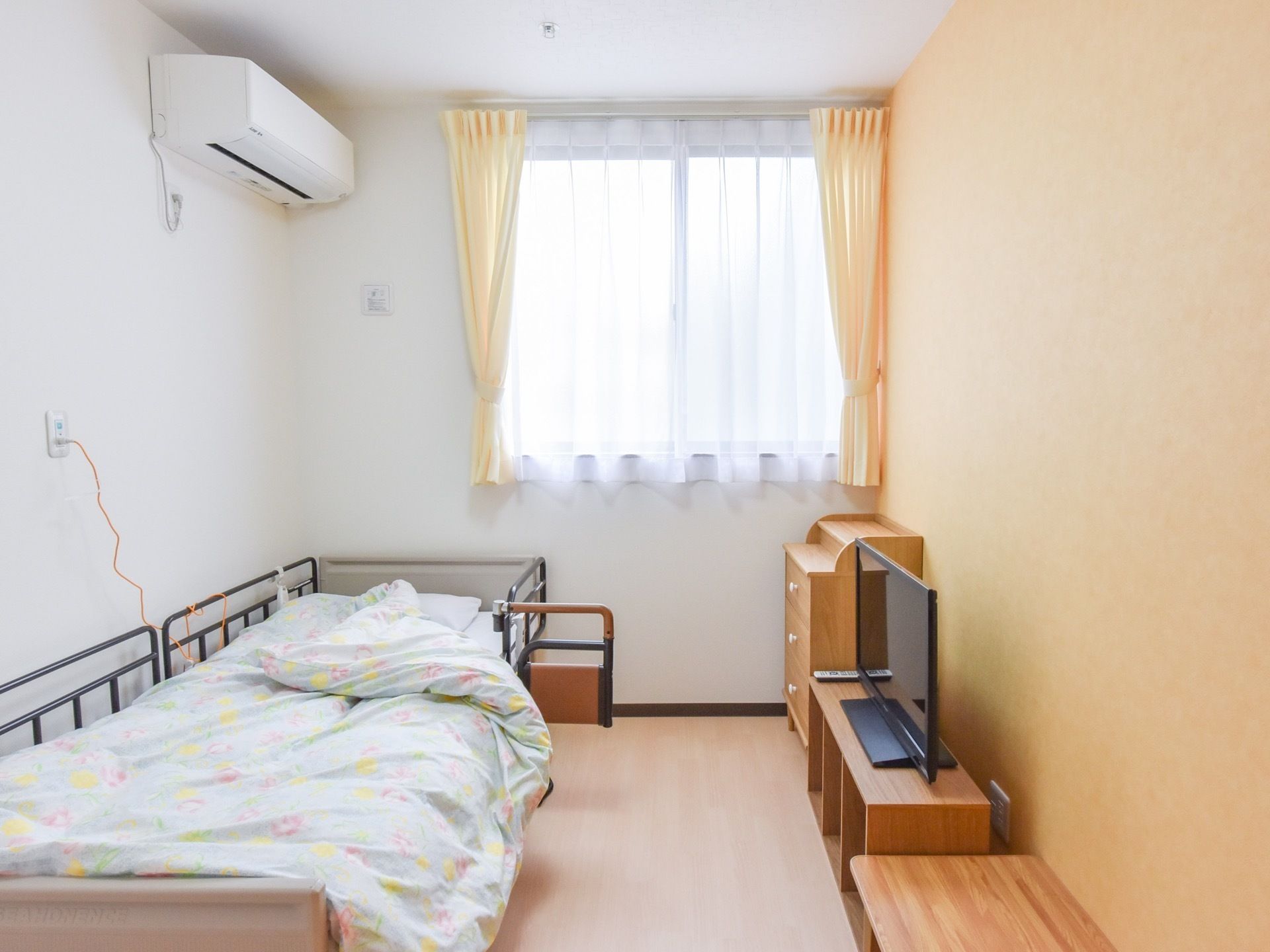 居室の一例。壁際に手すりのついたベッドが置かれている。小型のチェストと、テレビ台に液晶テレビが用意されている。