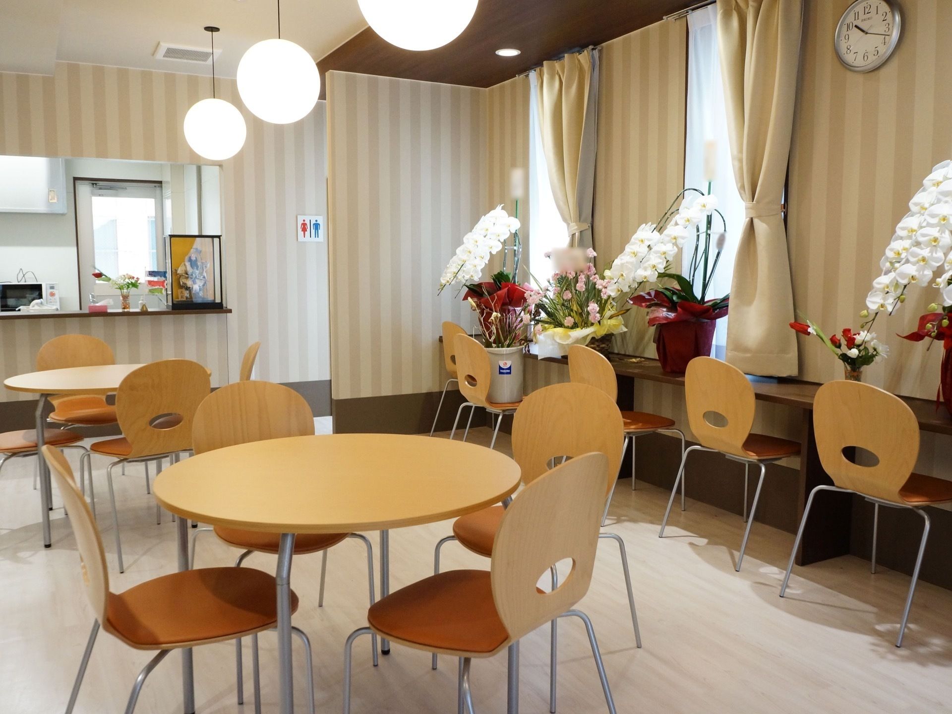 窓際には花が飾られている。４人掛けのテーブルと椅子がいくつか置かれている。壁紙はベージュ系と白系のストライプになっていてる。