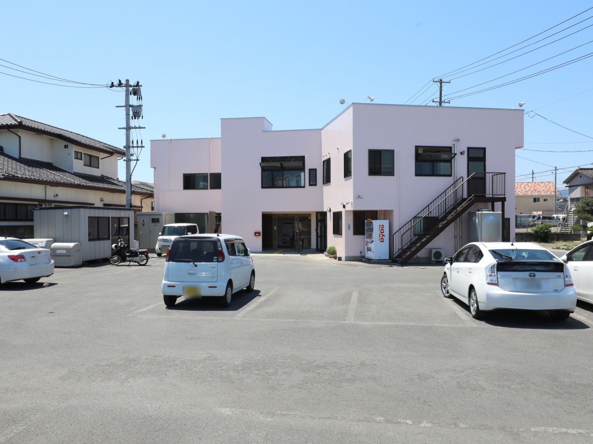 外壁は淡いピンク色で優しい雰囲気である。２階建ての建物で、建物の周囲には広い駐車場がある。入口近くには自動販売機が設置されている。