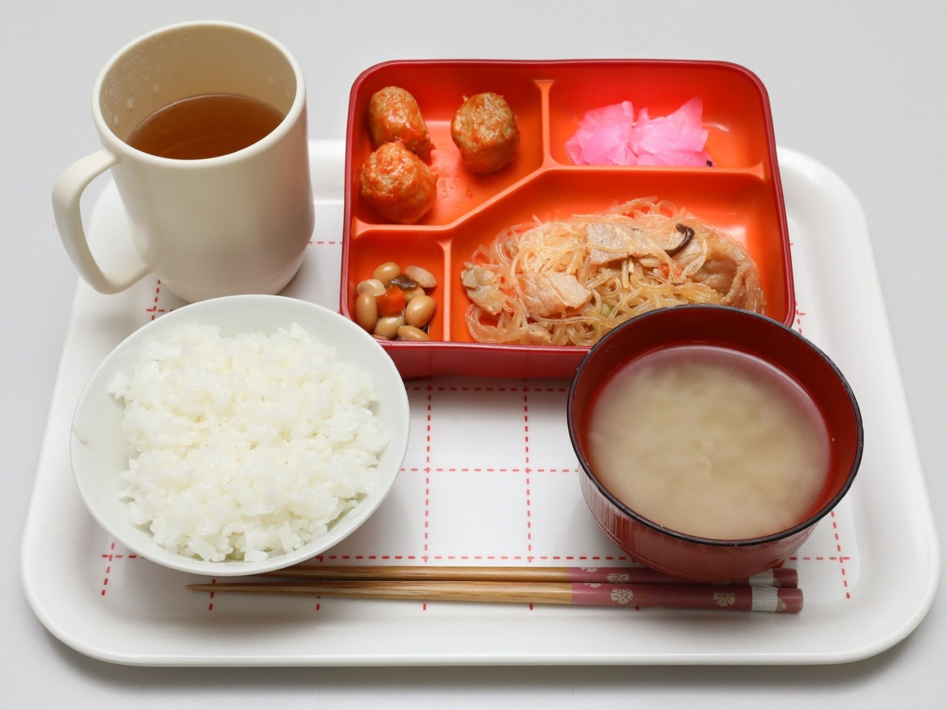 赤い区切りのあるプレートにおかずが４品盛り付けられている。ご飯と味噌汁、お茶と一緒にトレーに乗っている。