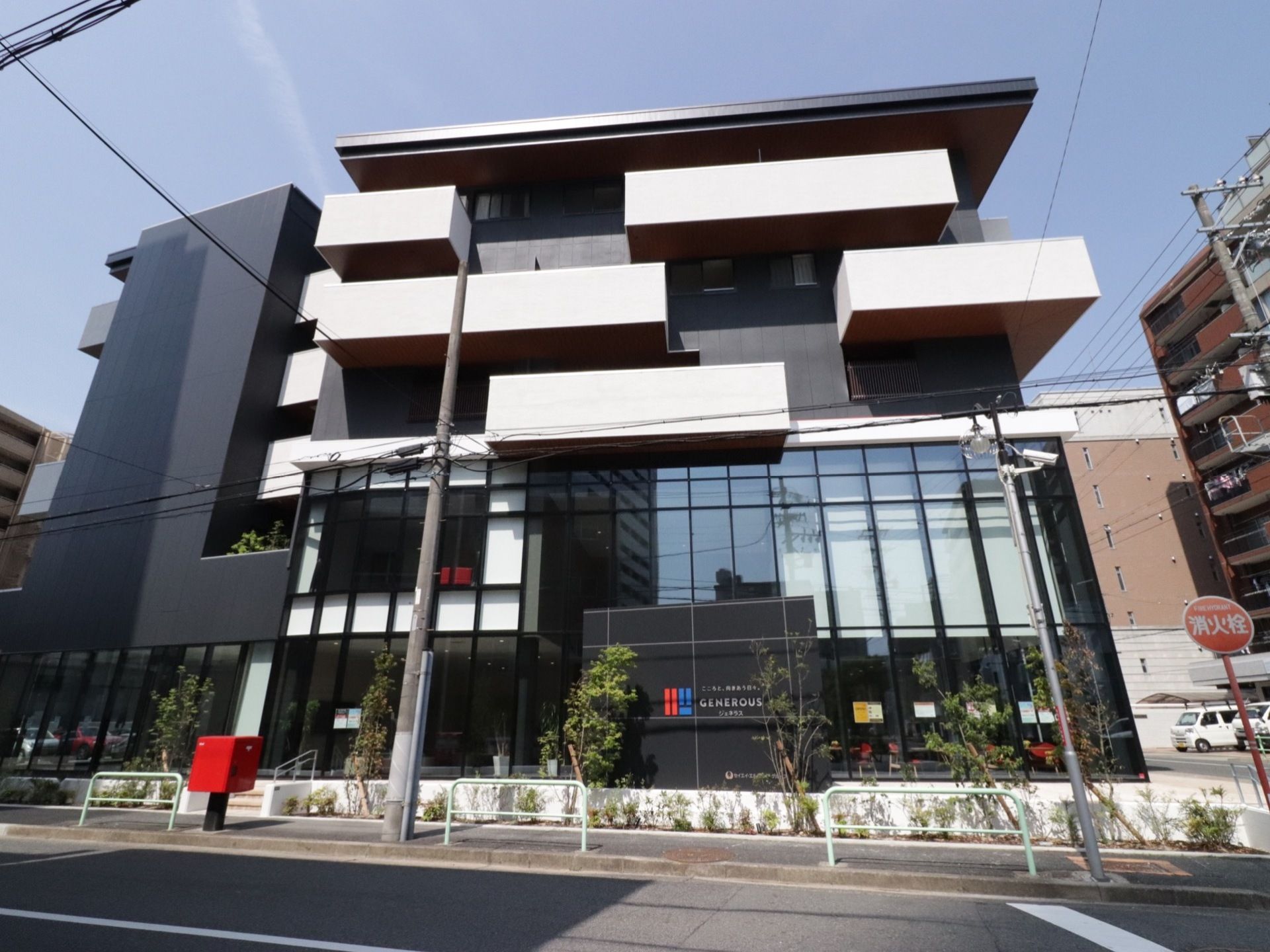 所々に凹凸のある建物は5階建てで、1～2階がガラス張りになっている。建物外の歩道沿いには植栽がなされている。