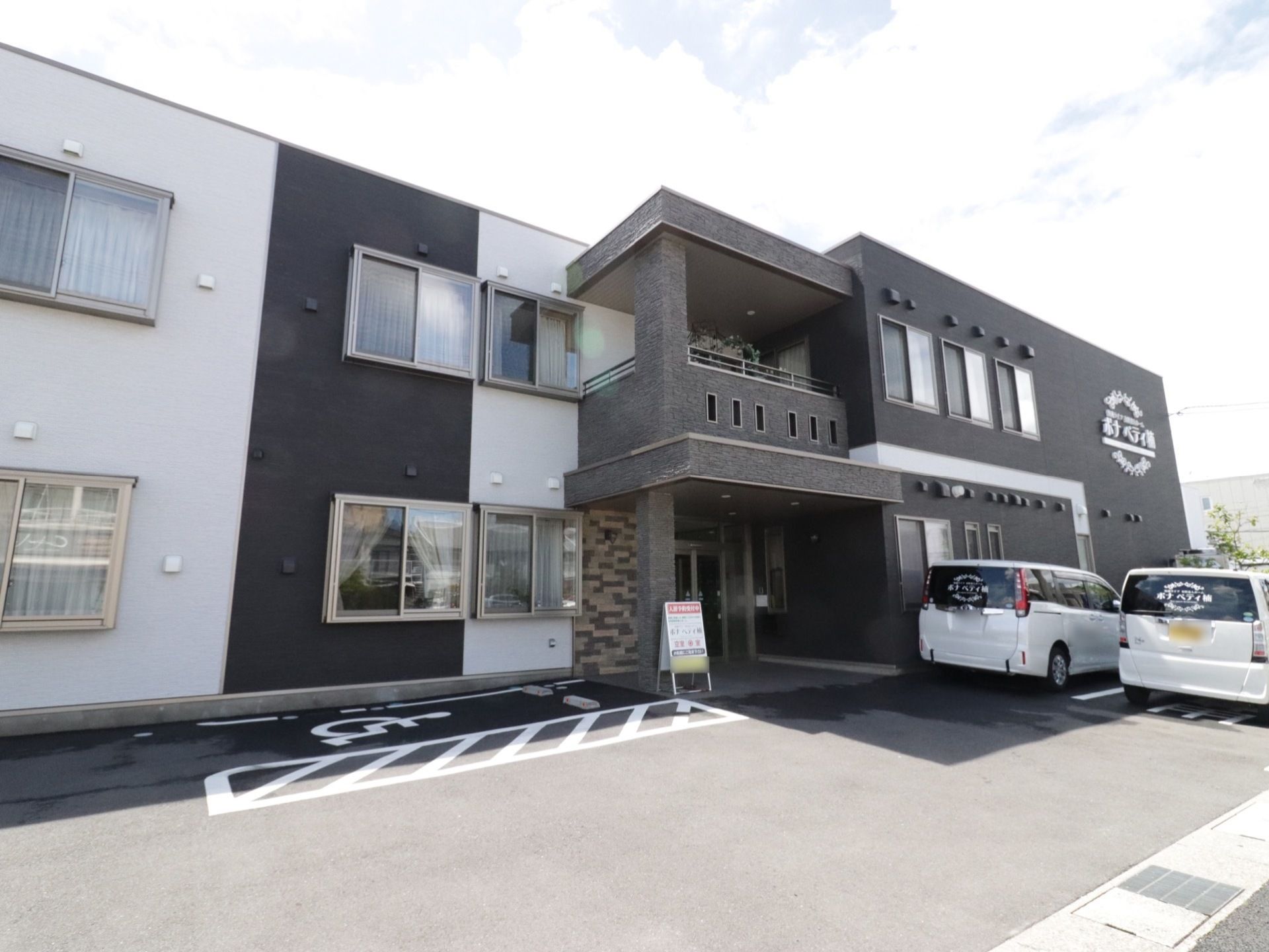 施設は、黒と白を基調としたスタイリッシュな外観となっている。玄関前には、駐車場スペースが確保されている。