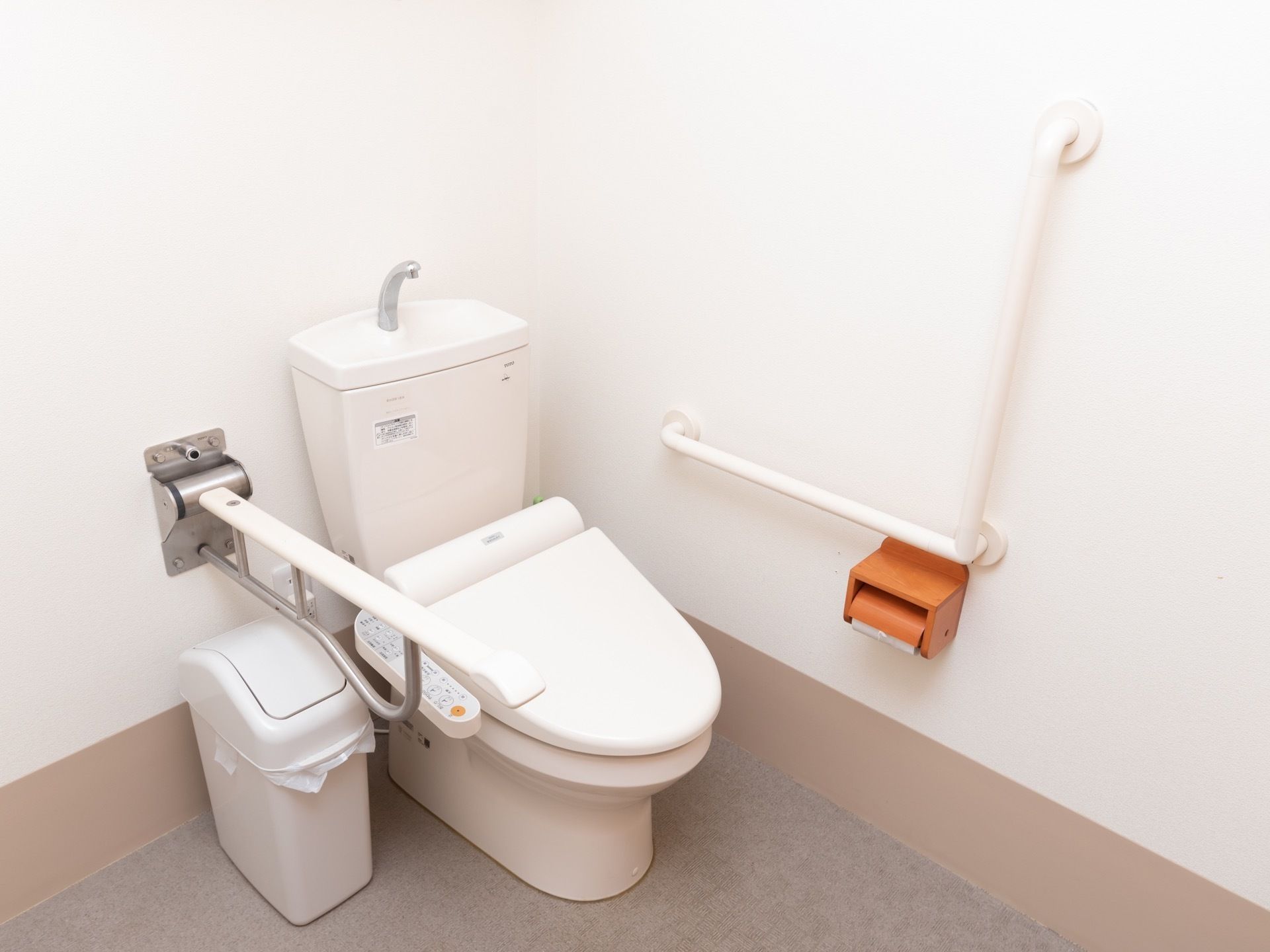 白を基調としたトイレである。壁にはL字型の手すりが取りつけられており、ペーパーホルダーが備わっている。