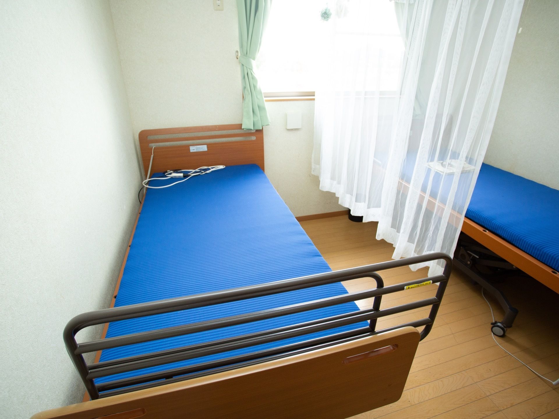 居室のベッドは青いマットが付いている。壁の中央に窓があり、緑色のカーテンをかけている。２つのベッドの間に、レースカーテンがある。