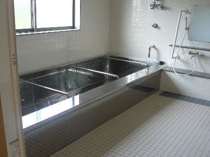 白いタイル壁に滑りにくい床の清潔感のある浴室。金属製のピカピカ光る浴槽には、横方向に手すりがわたされている。