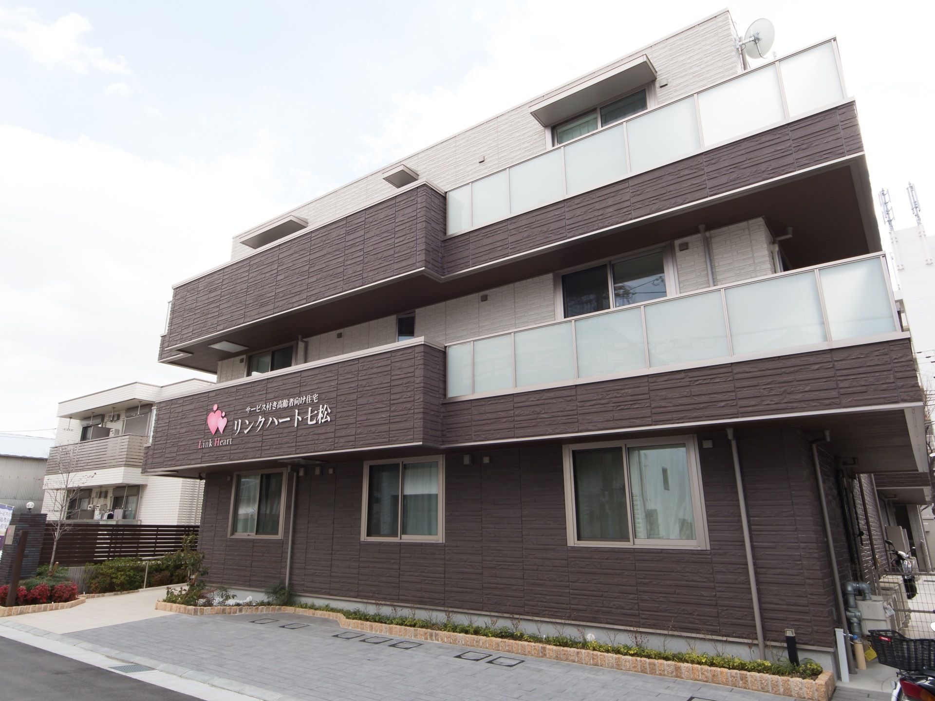 濃いブラウンとホワイトの壁が特徴的な外観をしている。建物の周りにはおしゃれな花壇があり、駐車スペースも用意されている。
