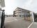 施設敷地の入口から見た画像である。黒い大きな門が開いており、奥に４階建ての施設が建っている。施設周囲は駐車場になっている。