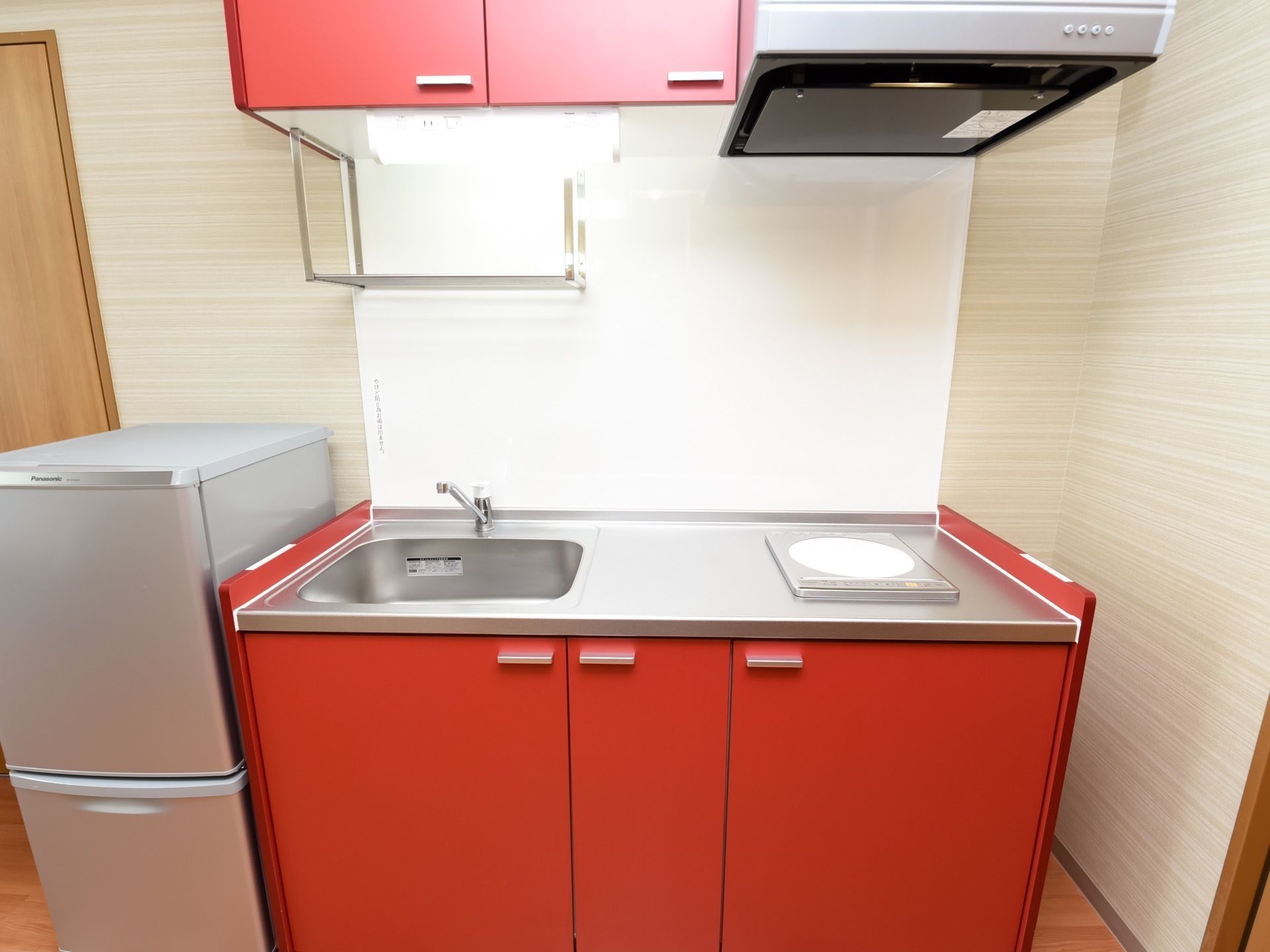 居室には、赤い収納扉が印象的な、IHコンロの付いたミニキッチンが完備されている。冷蔵庫が隣に置かれている。