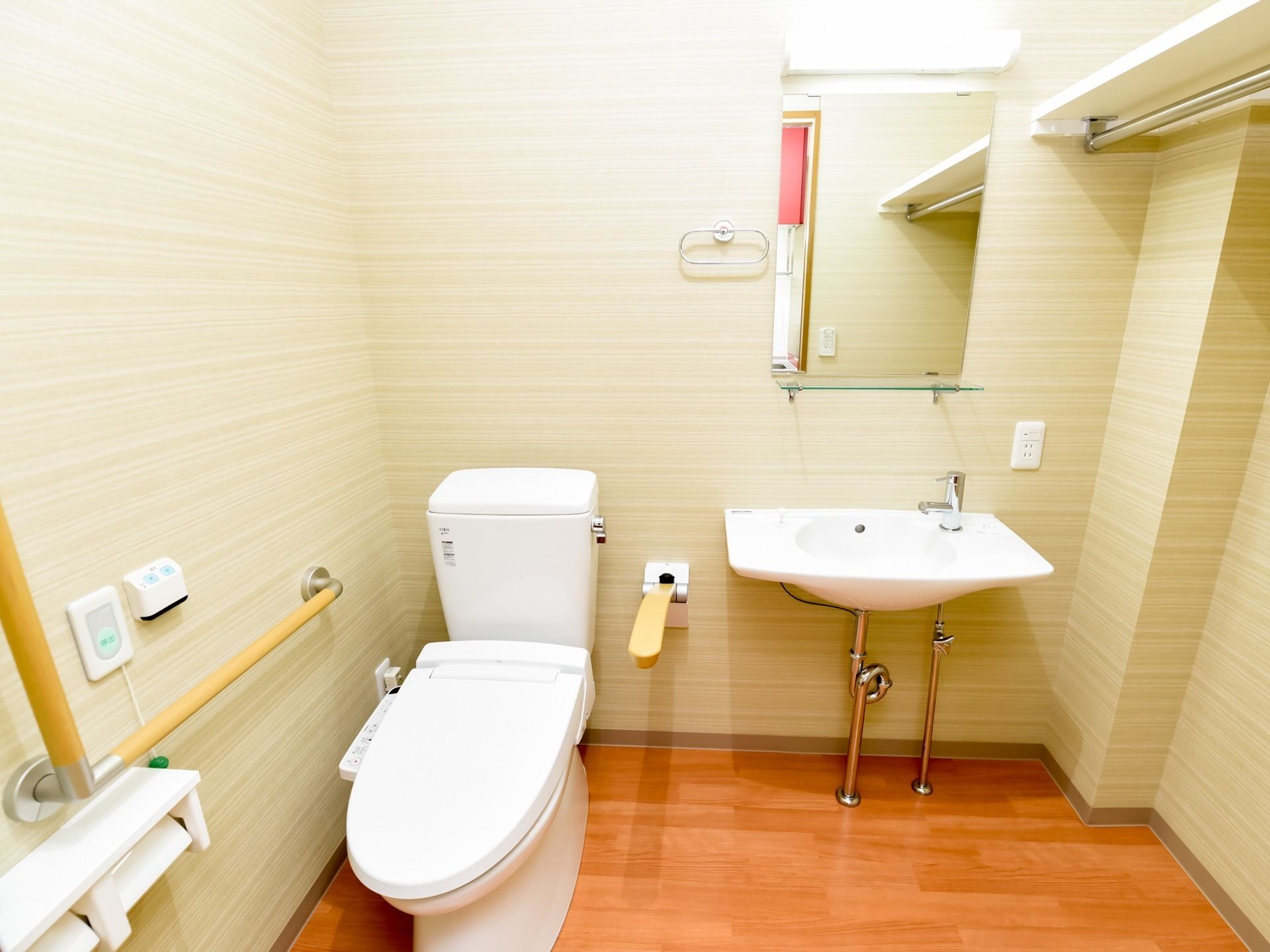 広いトイレには、洗面台や棚、手すりが完備されている。呼び出しボタンやタオル掛け、鏡が壁に取り付けられている。