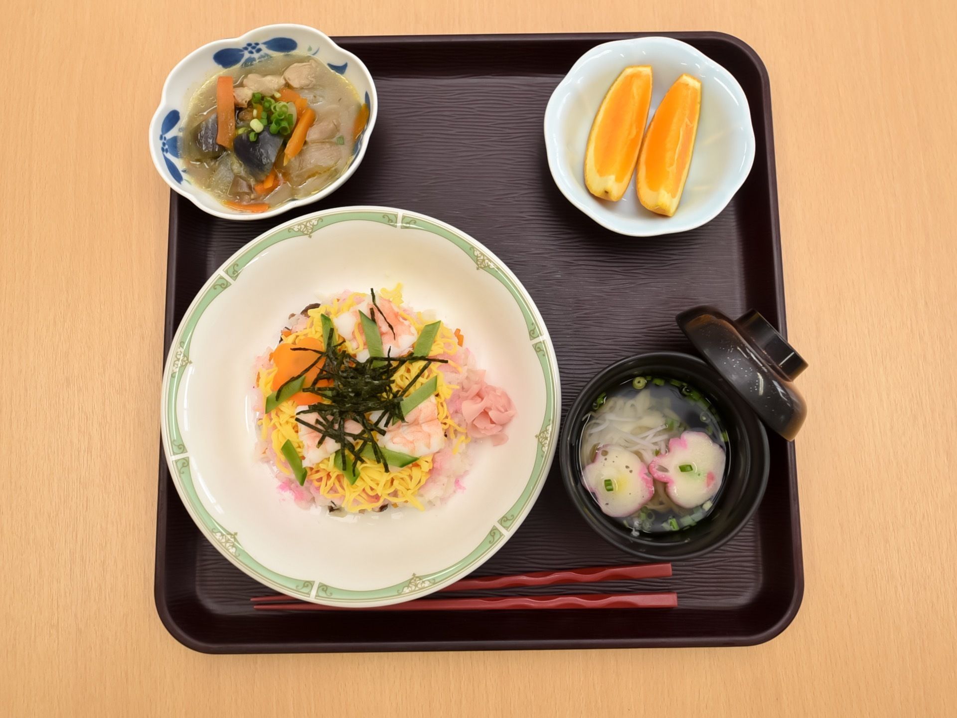 黒いトレイにちらし寿司とすまし汁、煮物が並べられており、カットオレンジが入った皿も置かれている。箸が用意されている。