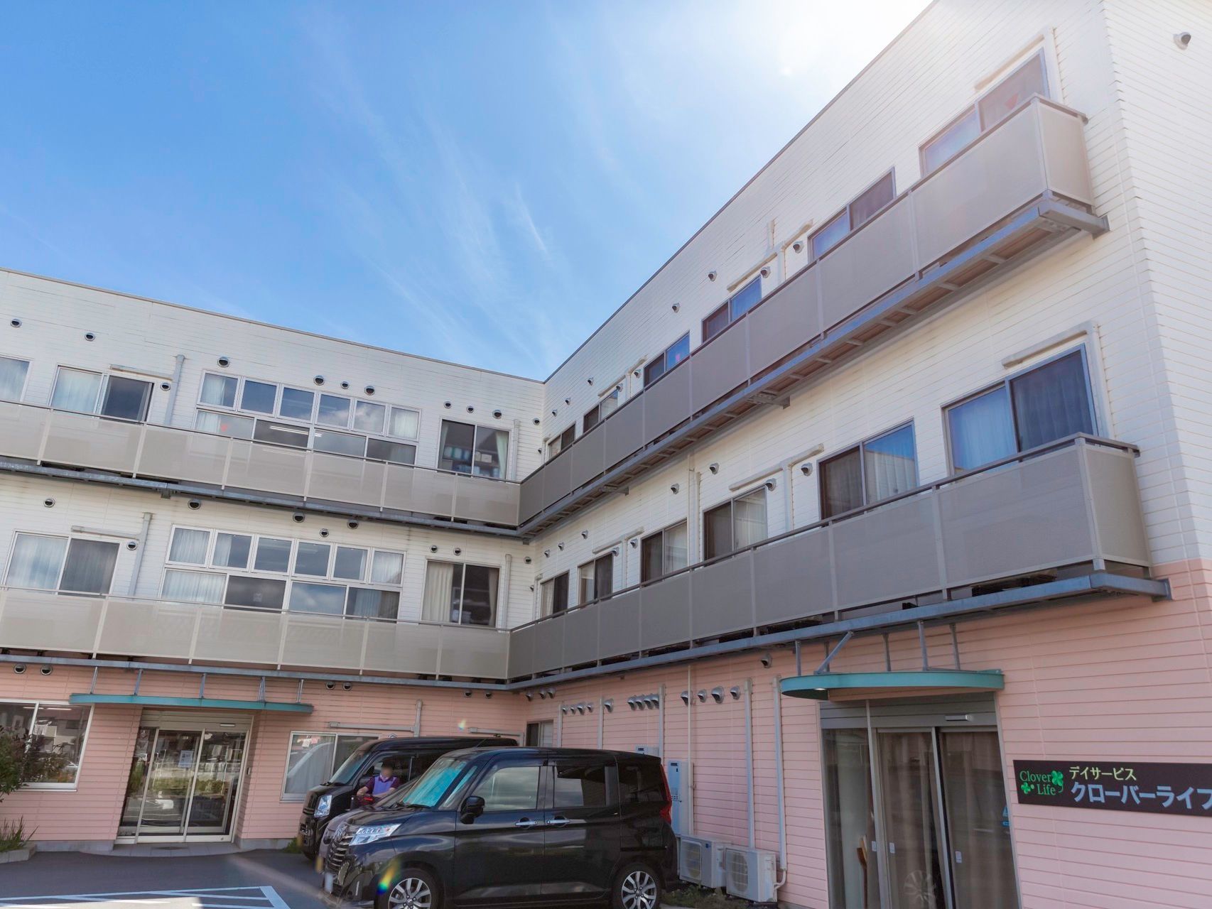 居室にベランダが備えつけられた、３階建ての施設である。親しみやすい色合いになっている。駐車場も完備されている。