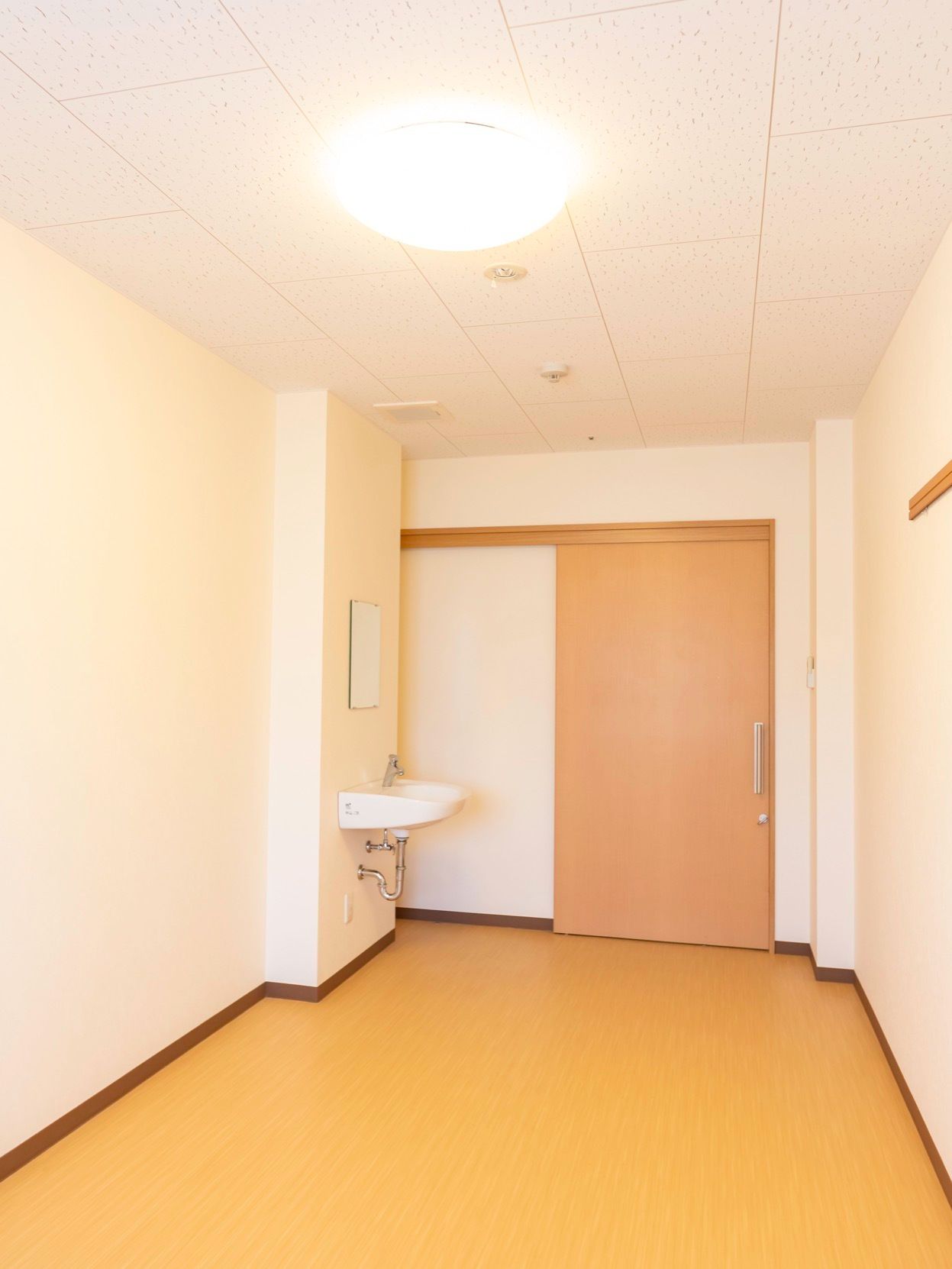 明るい色合いの居室を完備している。出入口のすぐそばには、鏡つきの洗面台がある。天井には丸形の照明が設置されている。