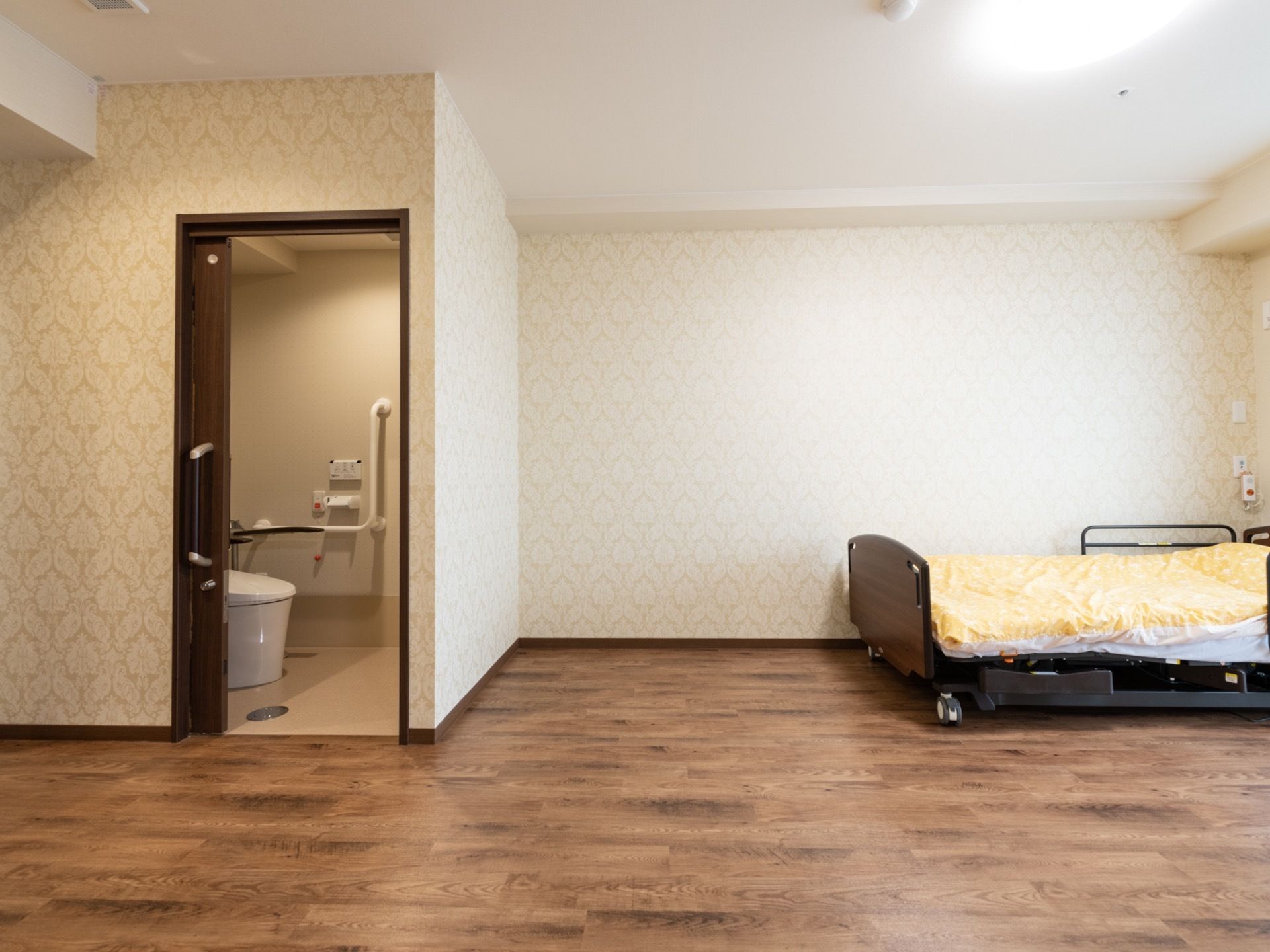 居室にはトイレがあり、壁際に介護用ベッドが置かれている。床はフローリングで白い壁にナースコールが設置されている。