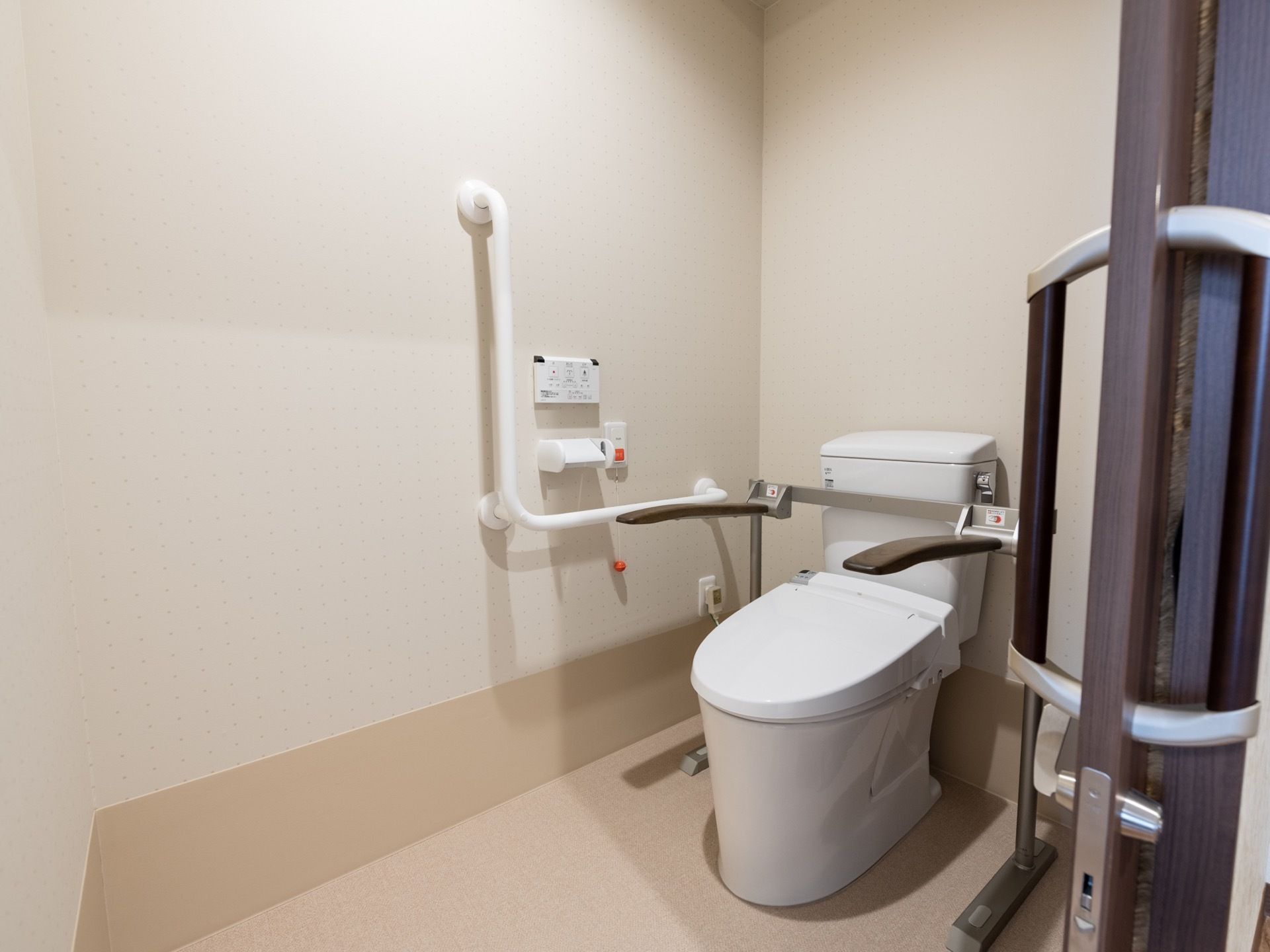 居室内のトイレは引き戸になっている。温水洗浄便座が取りつけられ、壁に操作用のパネルと、非常ボタンがある。