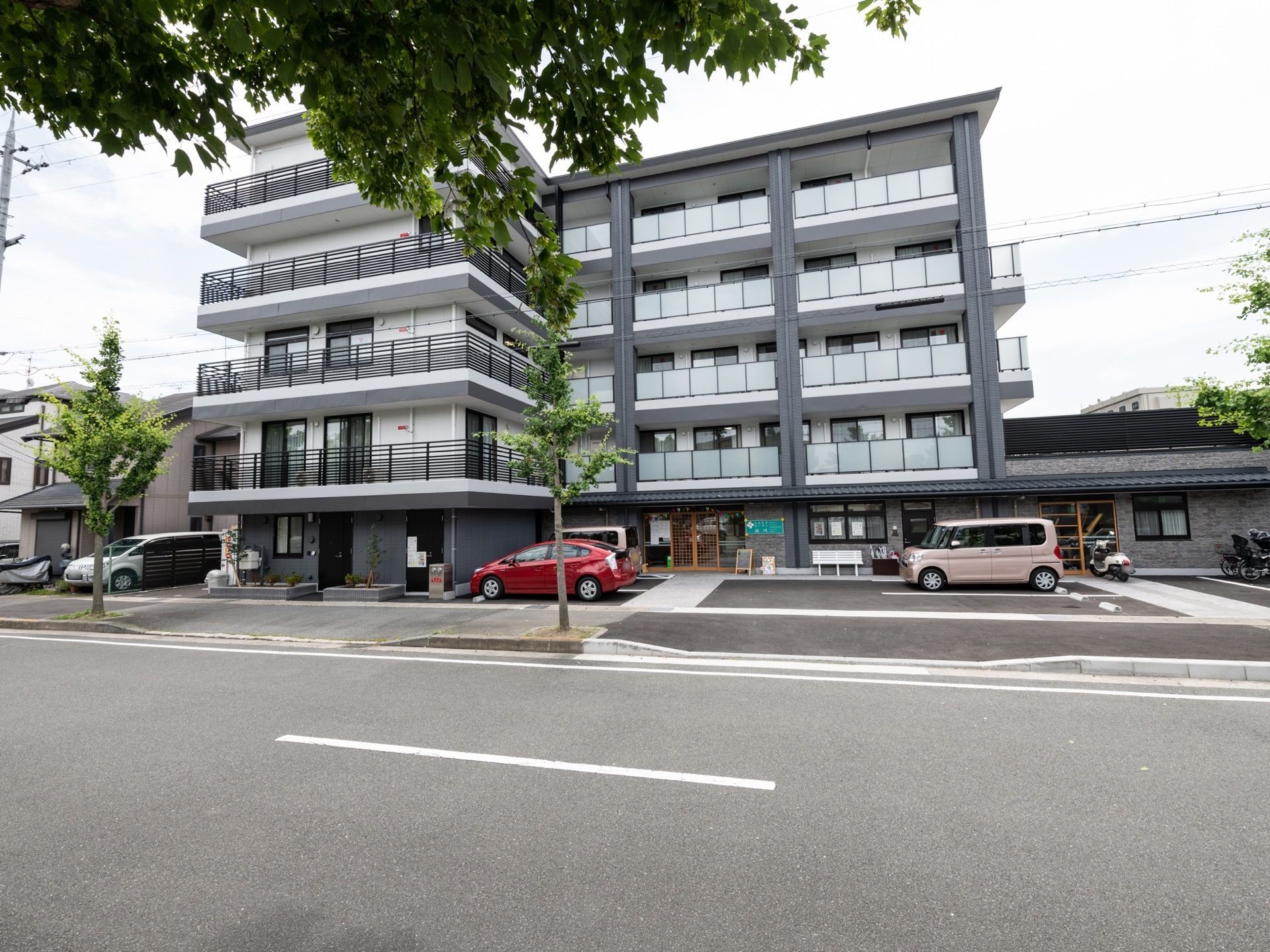 ５階建てのマンション風の建物で、ダークグレーの壁にアクリル板を設置したベランダが並んでいる。施設前は駐車スペースがある。