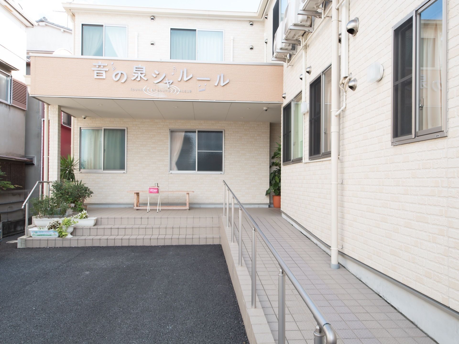 白を基調とした外壁の２階建ての建物である。鉢植えで緑を感じることができる。喫煙スペースが設けられている。