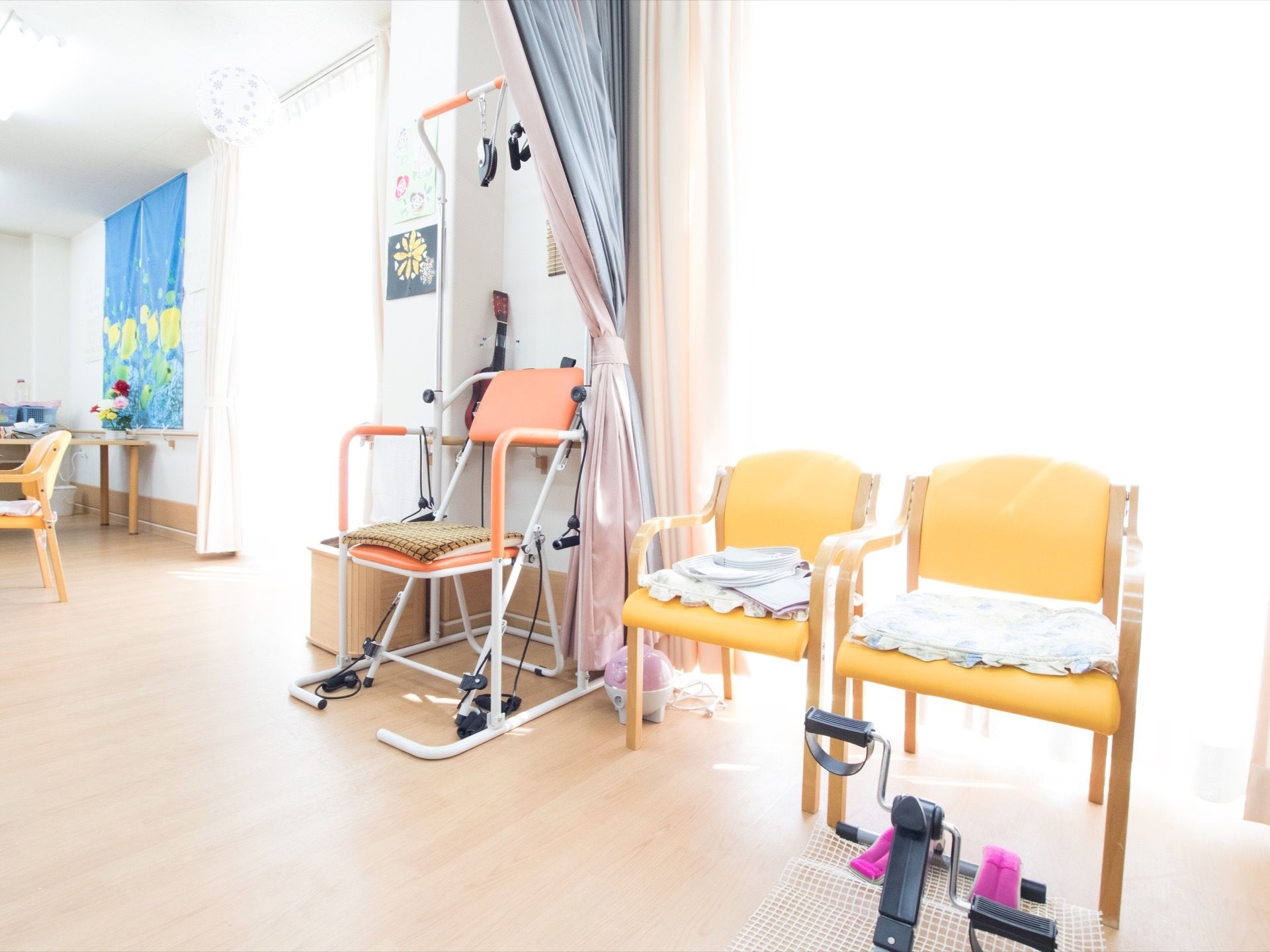 大きな掃き出し窓から日差しが差し込む、明るいフロア。窓際に椅子が並んでおり、足元にはペダル式のトレーニング器具が置かれている。