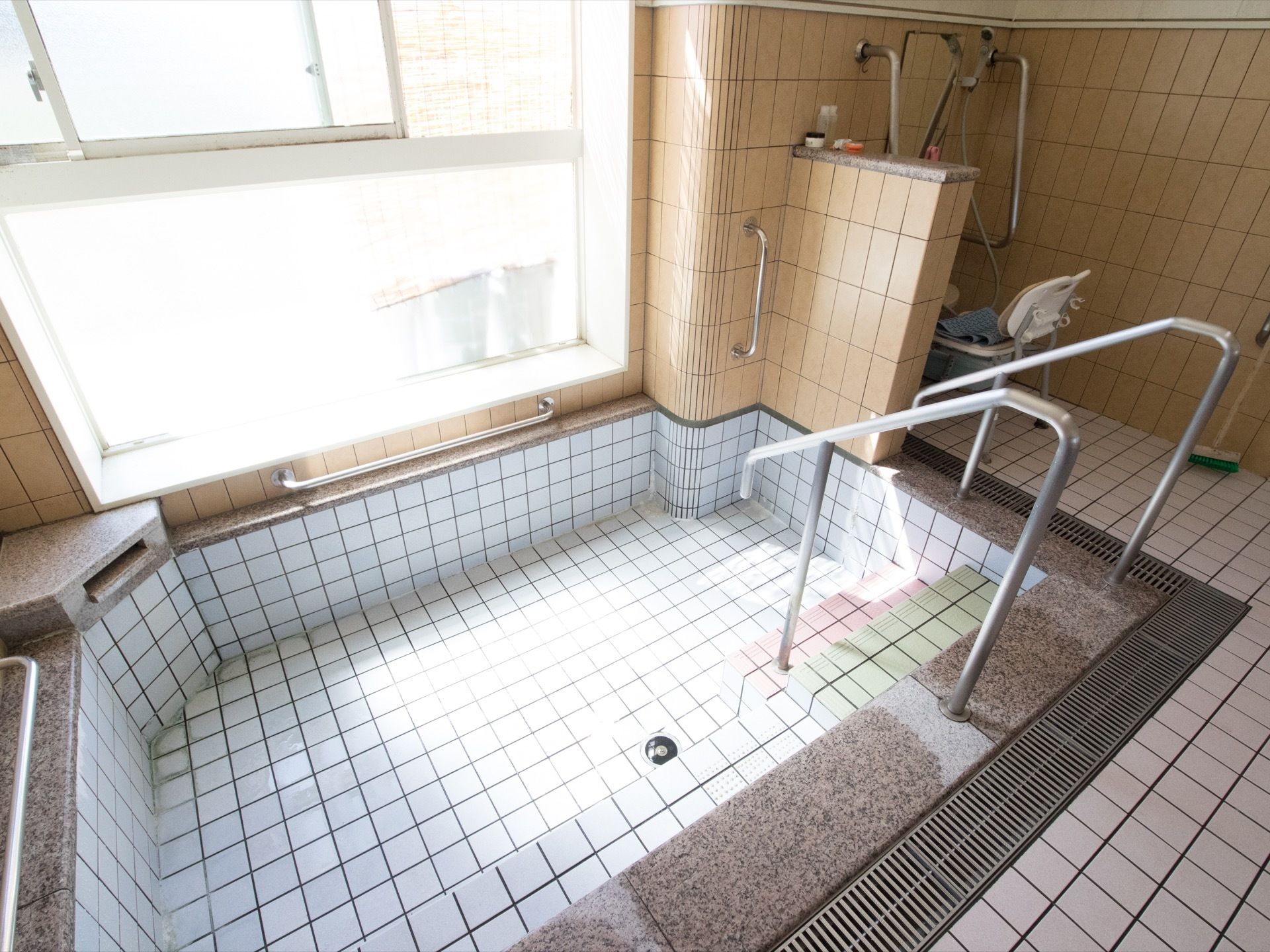 広々とした浴槽には安全面を考慮して、手すりのついたステップが設けられている。浴場奥のシャワー前には、シャワーチェアが置かれている。