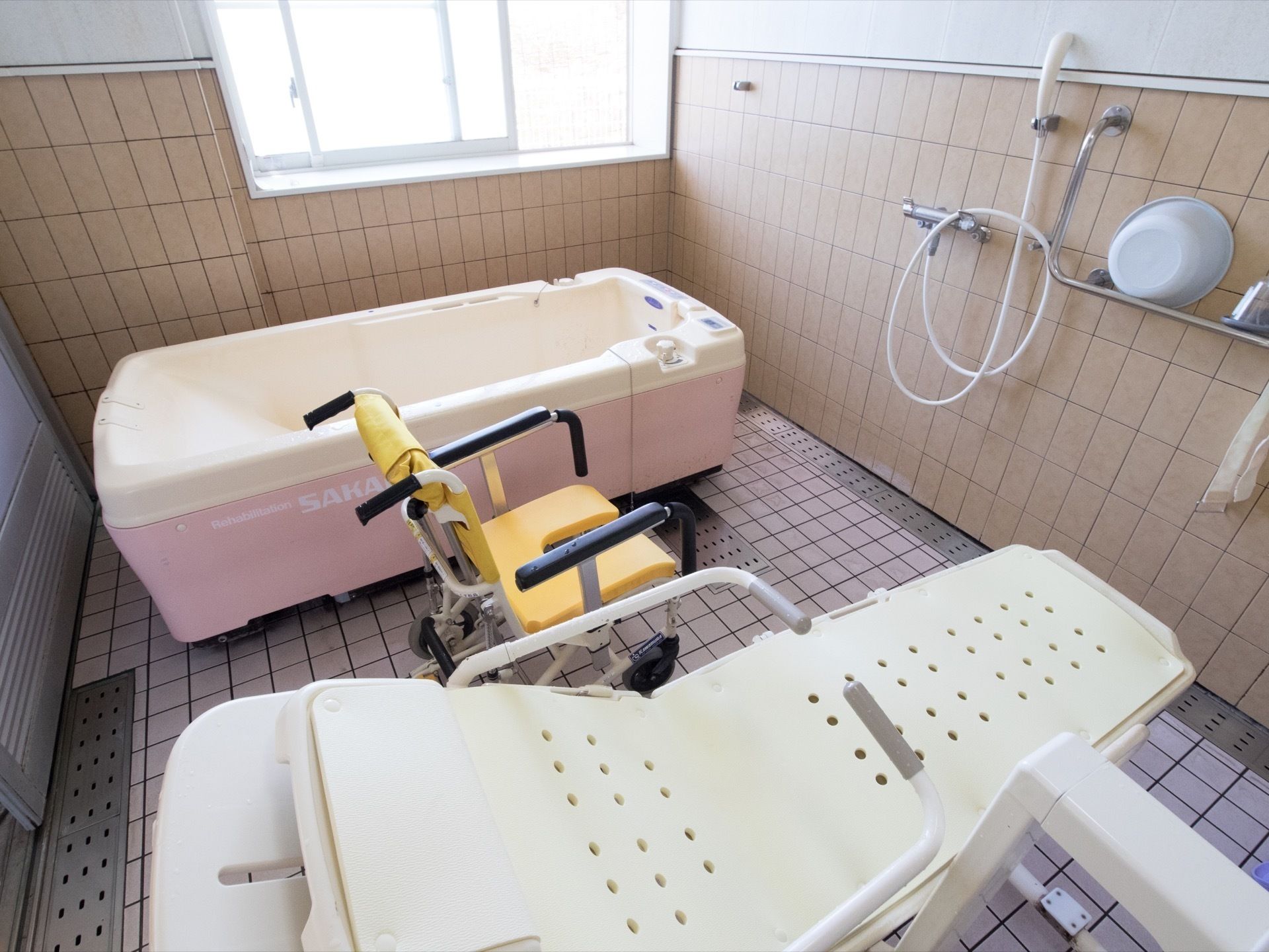入浴用の白いストレッチャーと黄色いシャワーチェア、ピンクの機械入浴機器が並んだ浴室